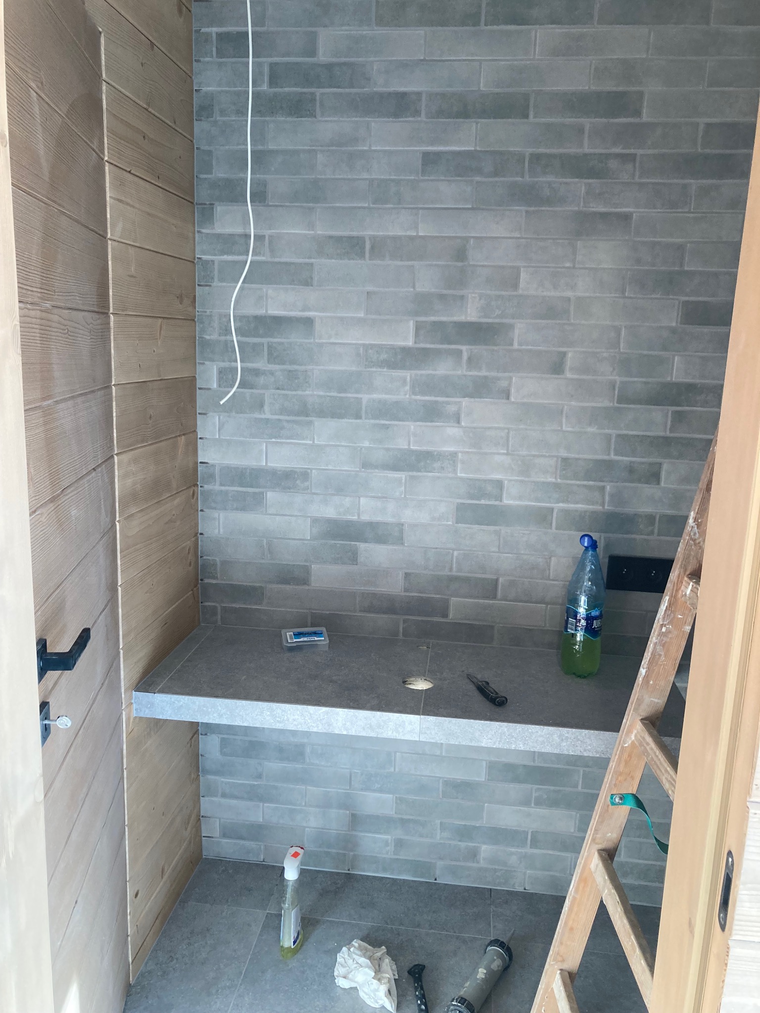 Wykończona ściana z szarych, prostokątnych płytek w łazience. Widoczny blat z otworem, drewniana boazeria i drabina. Na podłodze narzędzia i butelka z płynem.