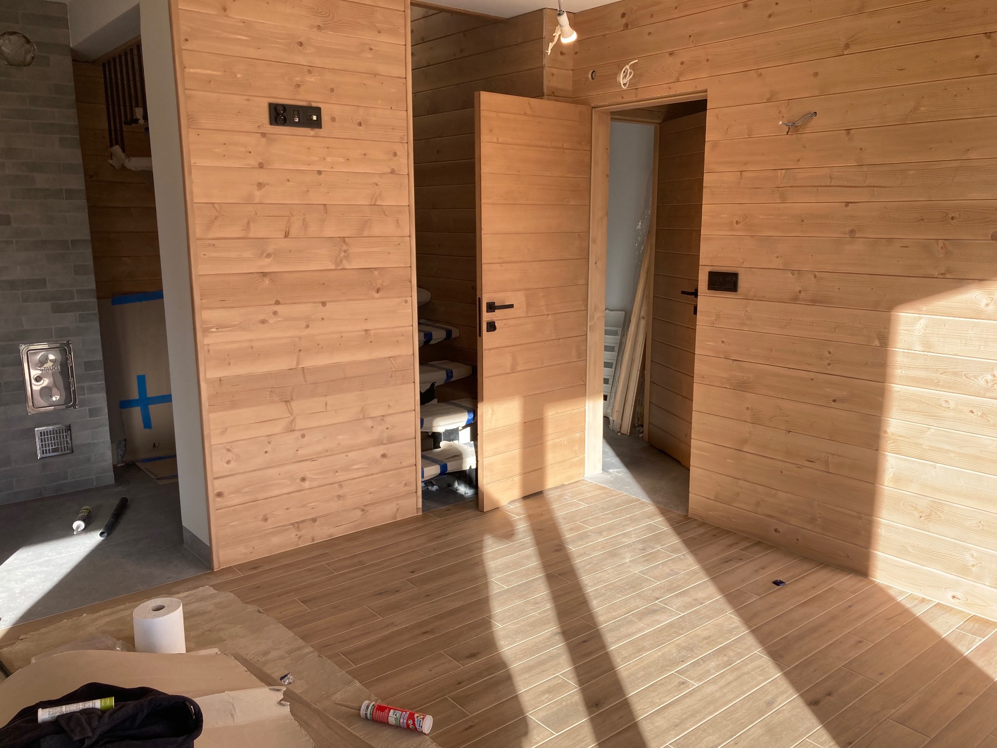 Wnętrze z wykończonymi drewnianymi ścianami i podłogą z paneli imitujących drewno. Widoczne otwarte drzwi i narzędzia, co sugeruje etap prac wykończeniowych w nowym lub remontowanym pomieszczeniu.