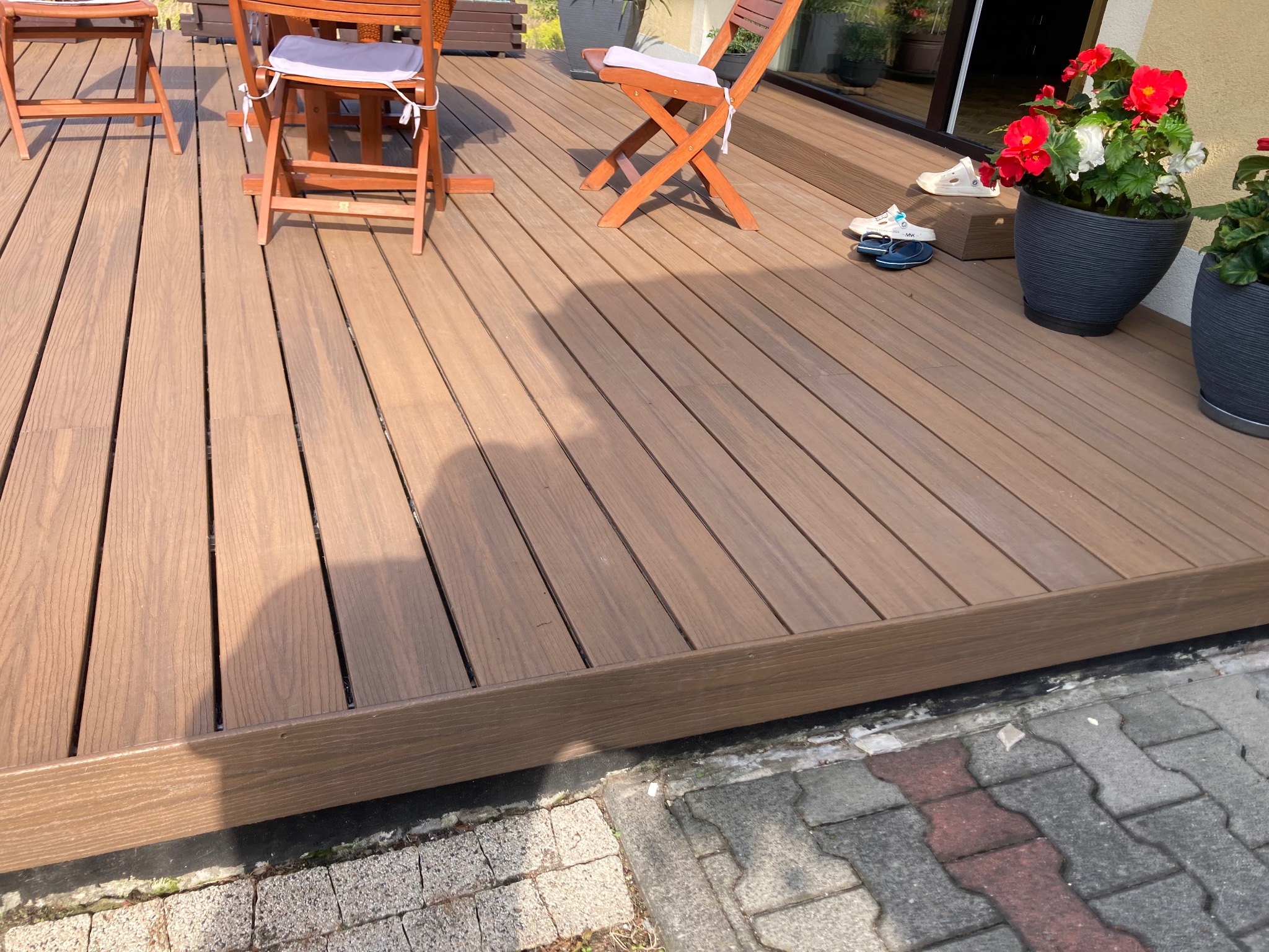 Nowoczesny taras z desek kompozytowych w odcieniach brązu, z drewnianymi krzesłami i donicami z kwiatami. Widoczne detale wykończenia i buty na tarasie.