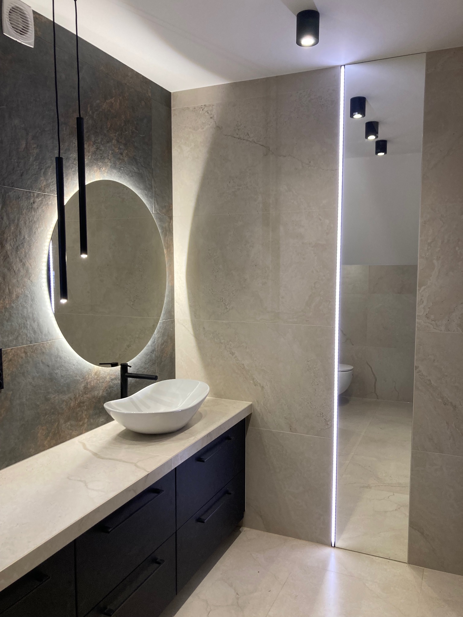 Nowoczesna łazienka z okrągłym lustrem LED, umywalką nablatową i ciemnymi szafkami. Jasne płytki imitujące kamień i oświetlenie punktowe tworzą elegancki klimat.