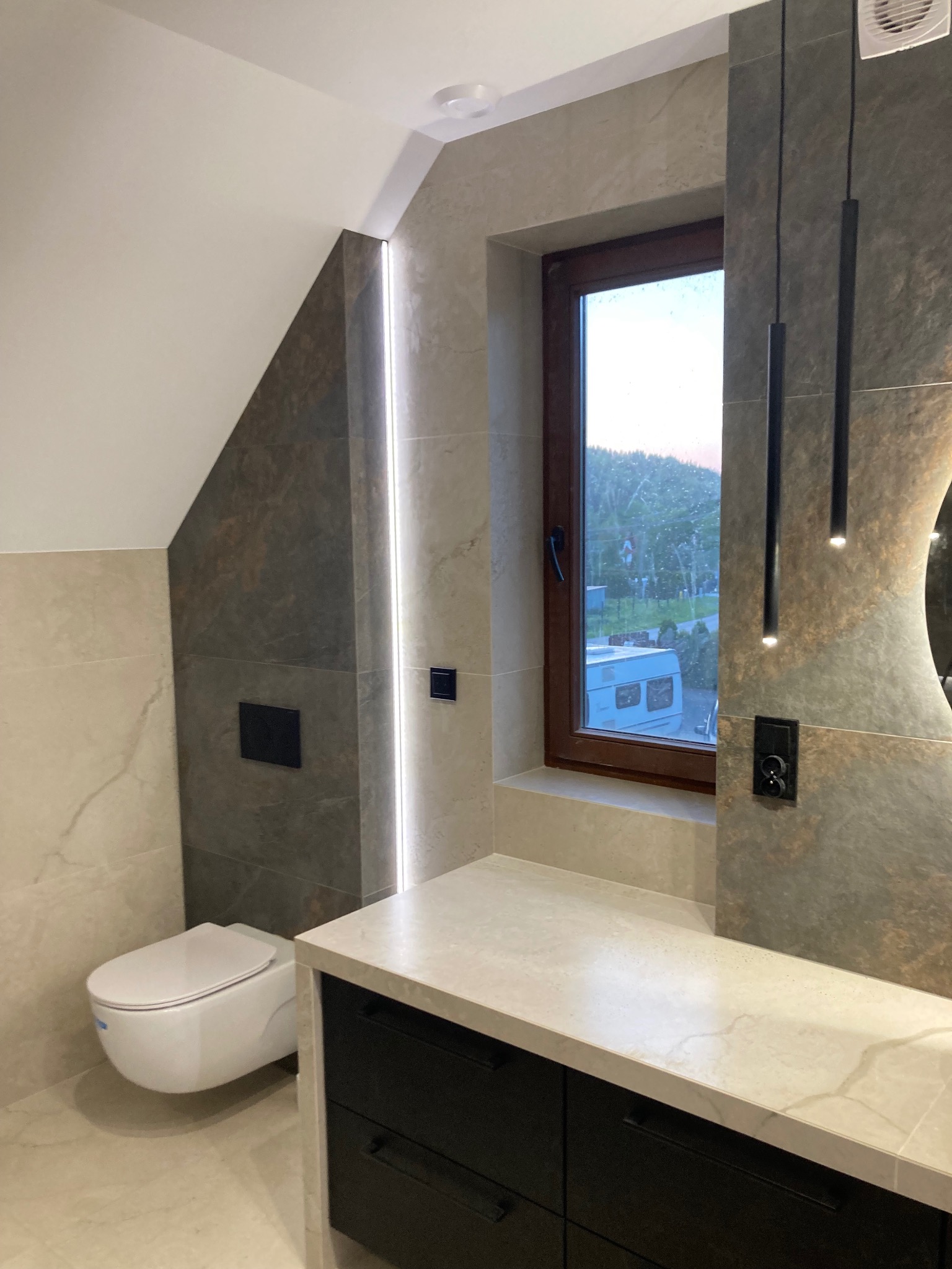 Nowoczesna łazienka na poddaszu z oknem, wykończona płytkami imitującymi kamień, podwieszana toaleta i czarna szafka pod umywalką z blatem.