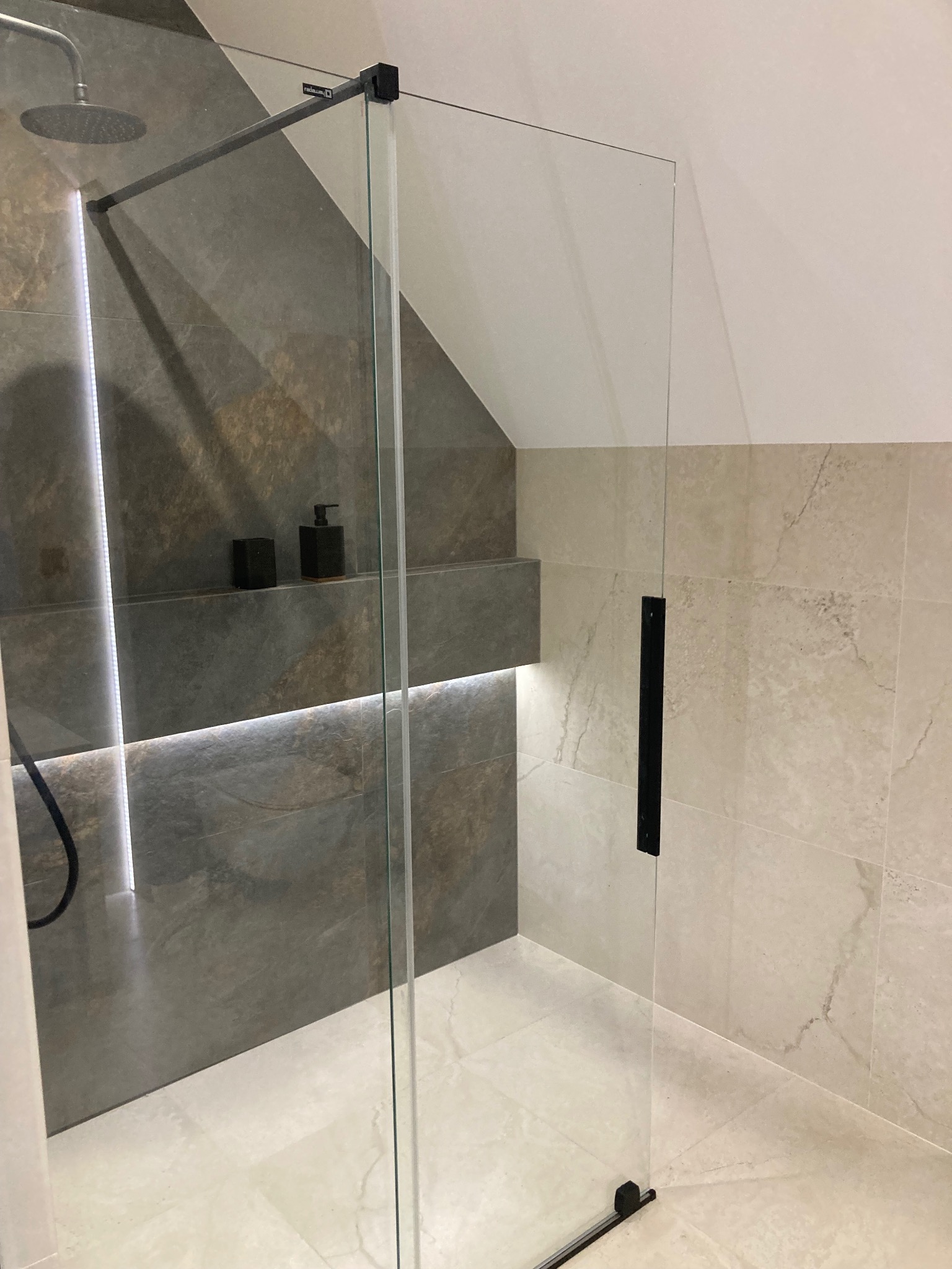 Kabina prysznicowa ze szklaną ścianką w łazience na poddaszu. Na ścianie półka z oświetleniem LED i kosmetykami. Płytki w odcieniach beżu i szarości.