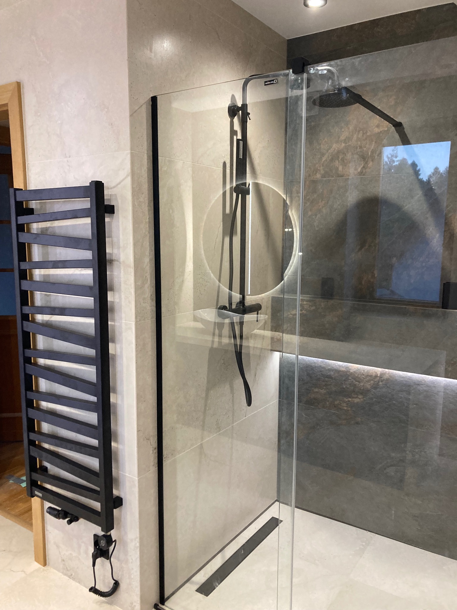 Nowoczesna kabina prysznicowa z czarnymi detalami, grzejnik drabinkowy i oświetlenie LED w wykończonej łazience z beżowymi i szarymi płytkami.