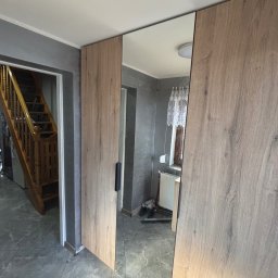 Tomasz Michalski - Szafa wnękowa z lustrem, drzwi przesuwne o teksturze drewna, szara ściana i podłoga, widoczny fragment schodów w tle. Minimalistyczny design.