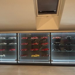 Tomasz Michalski - Trzy przeszklone szafy z podświetleniem LED, prezentujące kolekcję modeli samochodów w skali, ustawione na tle skośnego sufitu z oknem dachowym.