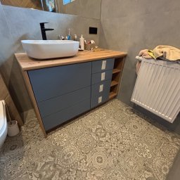 Tomasz Michalski - Nowoczesna szafka łazienkowa z drewnianym blatem i szarymi frontami szuflad. Umywalka nablatowa, czarna bateria, dekoracyjna podłoga. Widok na grzejnik i fragment ściany z lustrem.