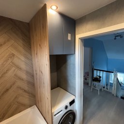 Tomasz Michalski - Łazienka z zabudową meblową: szafka nad pralką, drewniana obudowa wanny, płytki imitujące drewno na ścianie. Widok na korytarz z zabawkami w tle.