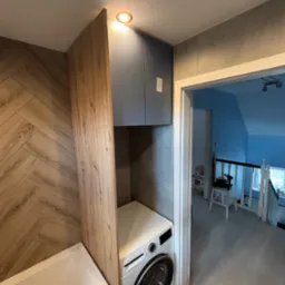 Łazienka z zabudową meblową: szafka nad pralką, drewniana obudowa wanny, płytki imitujące drewno na ścianie. Widok na korytarz z zabawkami w tle.