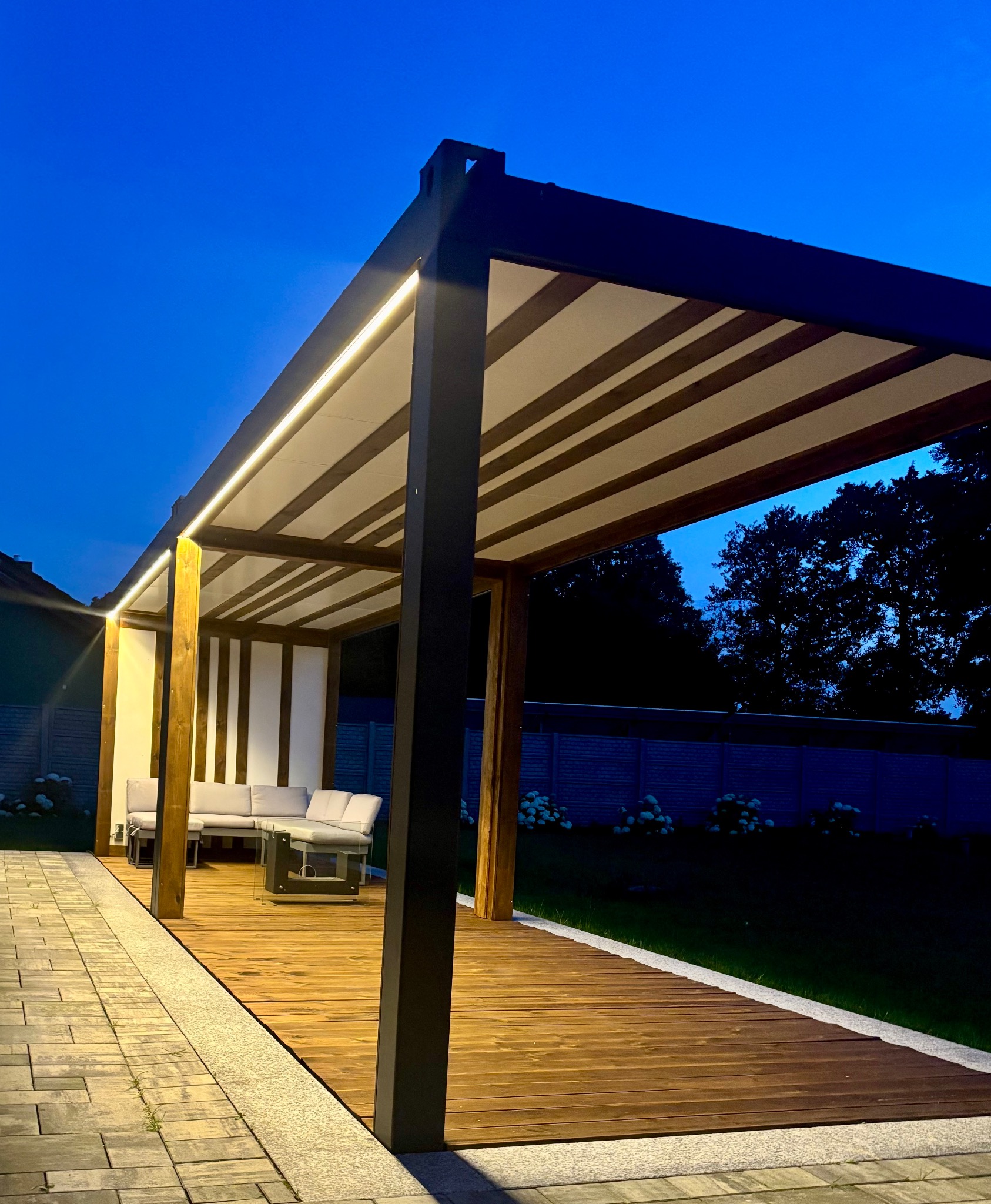 Nowoczesna pergola z oświetleniem LED i drewnianym tarasem, z meblami ogrodowymi, sfotografowana o zmierzchu na tle zielonego ogrodu i niebieskiego nieba.