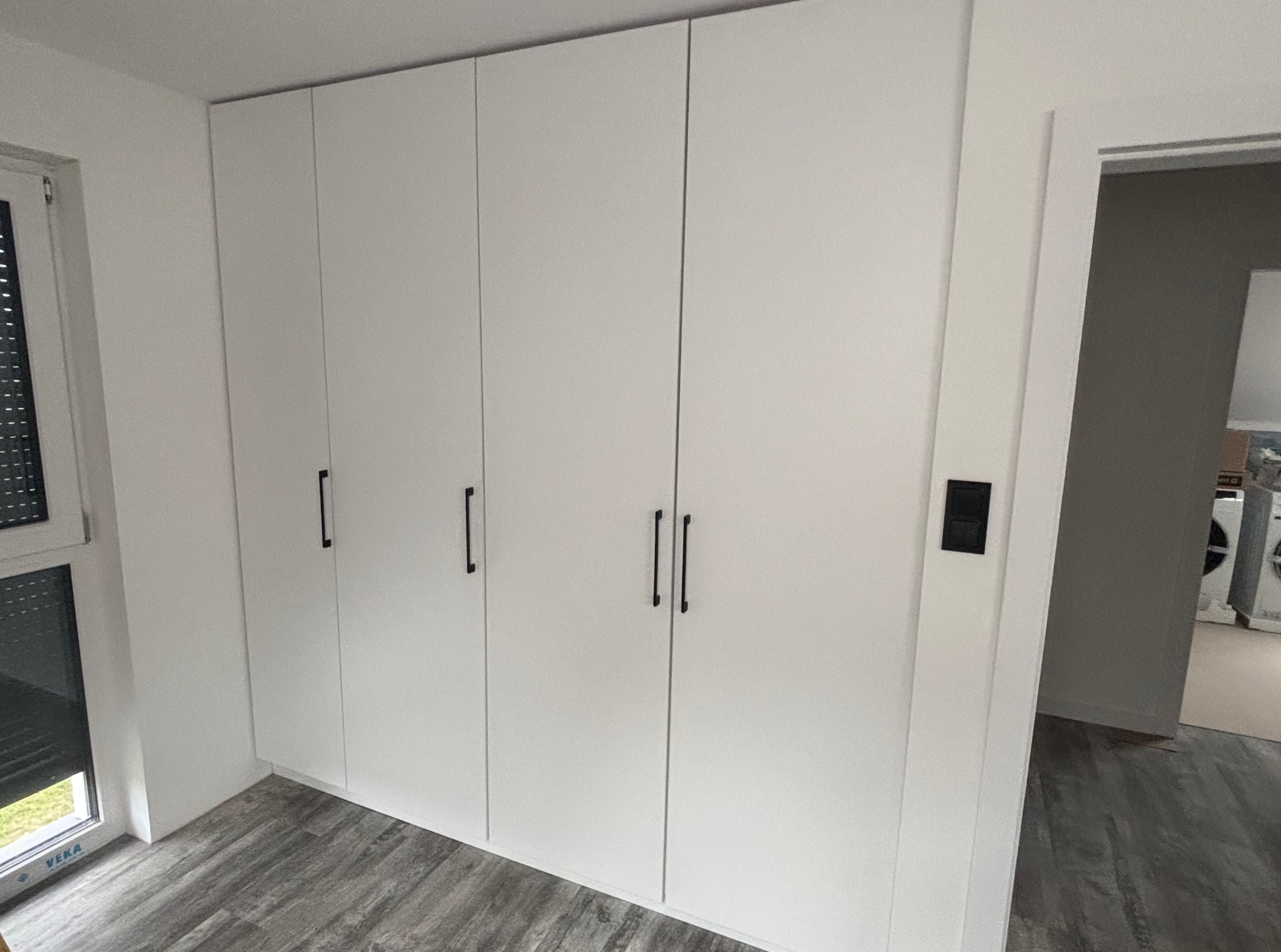 Biała szafa wnękowa z czarnymi uchwytami, widok z boku, na podłodze panele imitujące drewno, obok okno z żaluzjami i widok na wnętrze z pralkami.