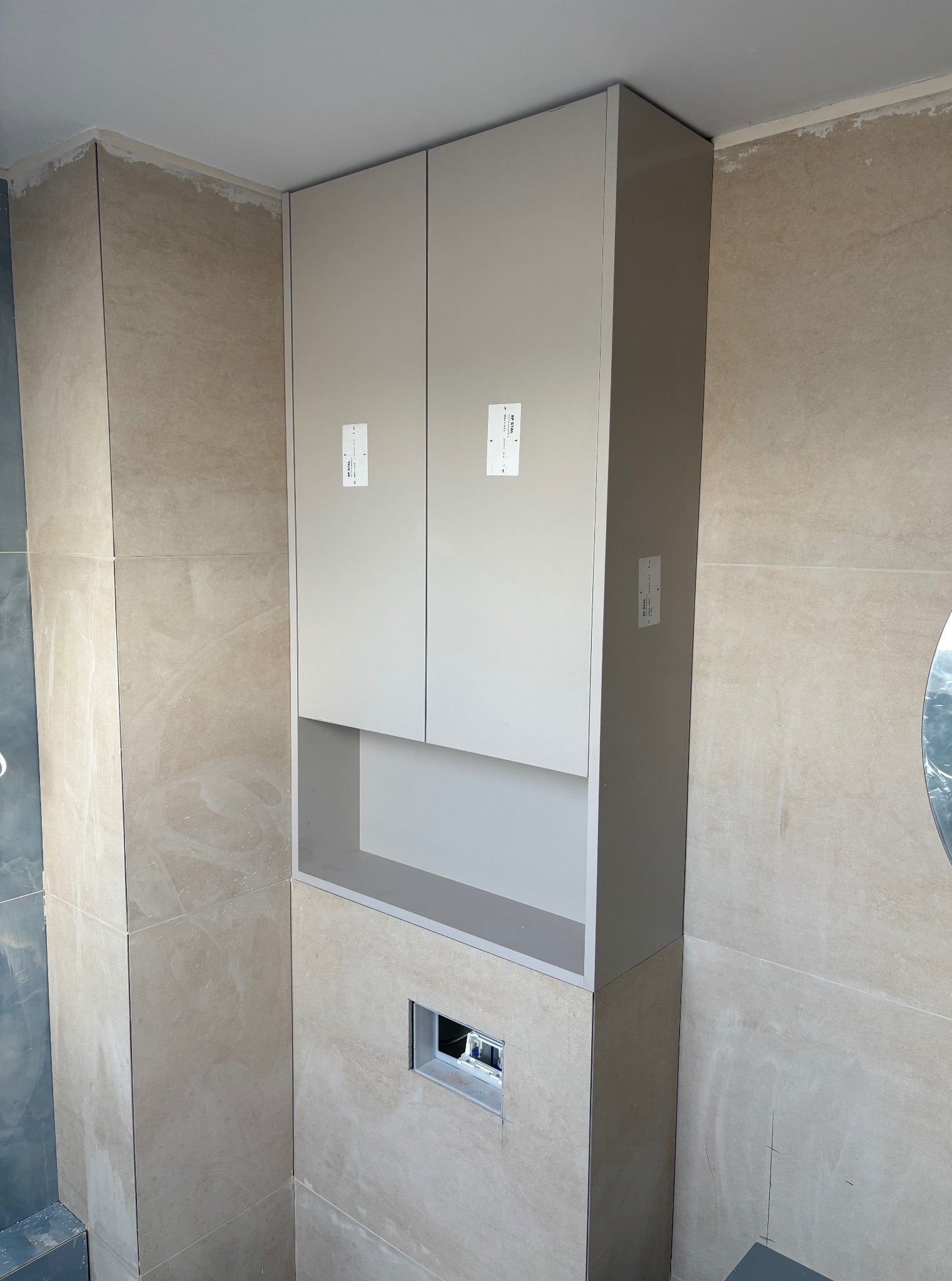 Pionowa szafka łazienkowa w odcieniach beżu, z otwartą półką nad zabudową stelaża WC. Widoczne płytki ścienne w kolorze piaskowym i sufit. Minimalistyczny design.