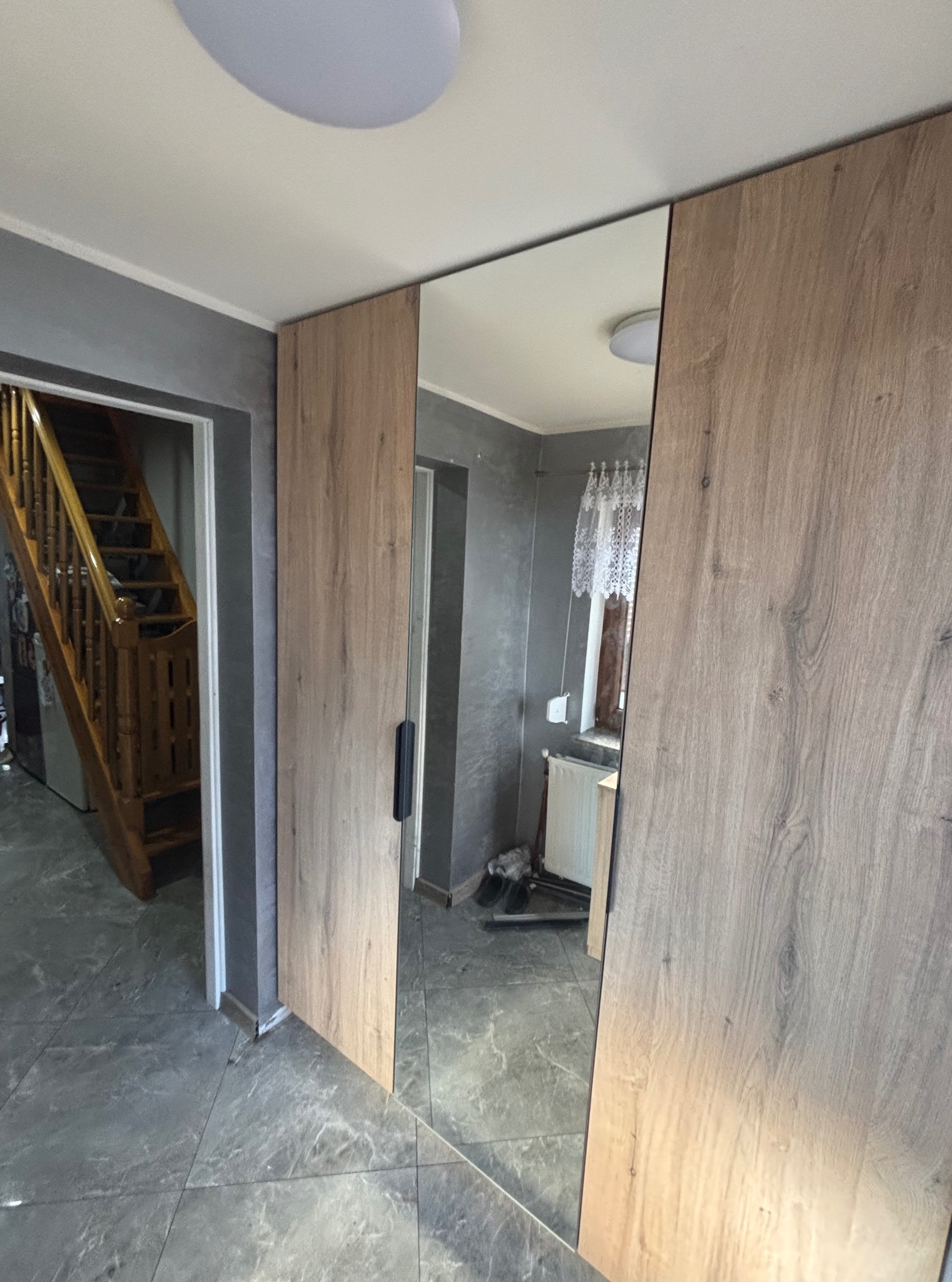 Szafa wnękowa z lustrem, drzwi przesuwne o teksturze drewna, szara ściana i podłoga, widoczny fragment schodów w tle. Minimalistyczny design.