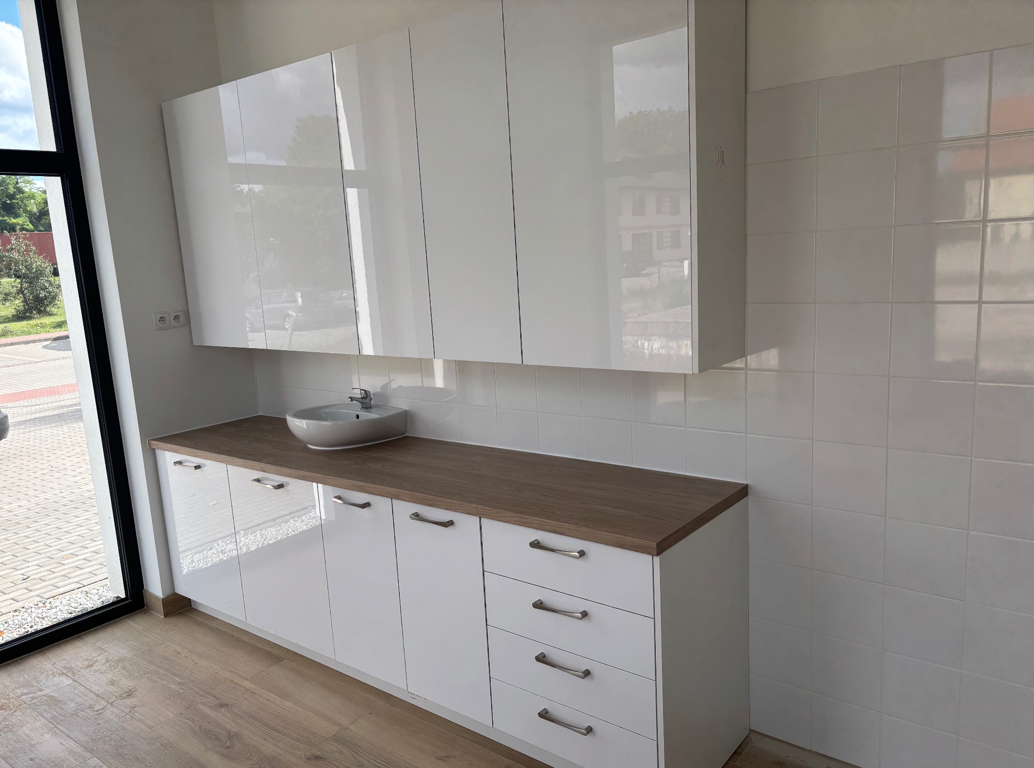 Białe, lakierowane szafki łazienkowe z drewnianym blatem i okrągłą umywalką, z widocznym oknem po lewej stronie i białymi kafelkami na ścianie.