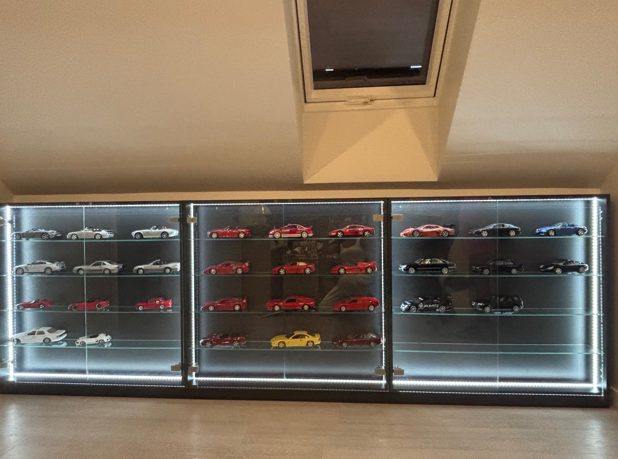 Trzy przeszklone szafy z podświetleniem LED, prezentujące kolekcję modeli samochodów w skali, ustawione na tle skośnego sufitu z oknem dachowym.