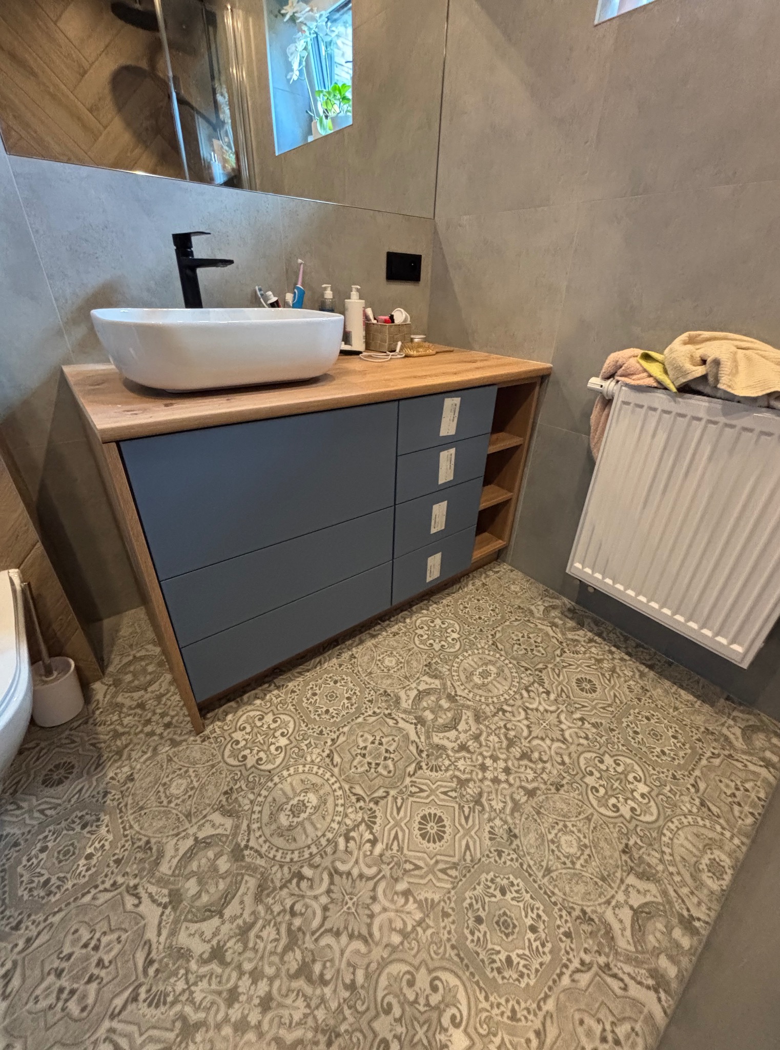 Nowoczesna szafka łazienkowa z drewnianym blatem i szarymi frontami szuflad. Umywalka nablatowa, czarna bateria, dekoracyjna podłoga. Widok na grzejnik i fragment ściany z lustrem.