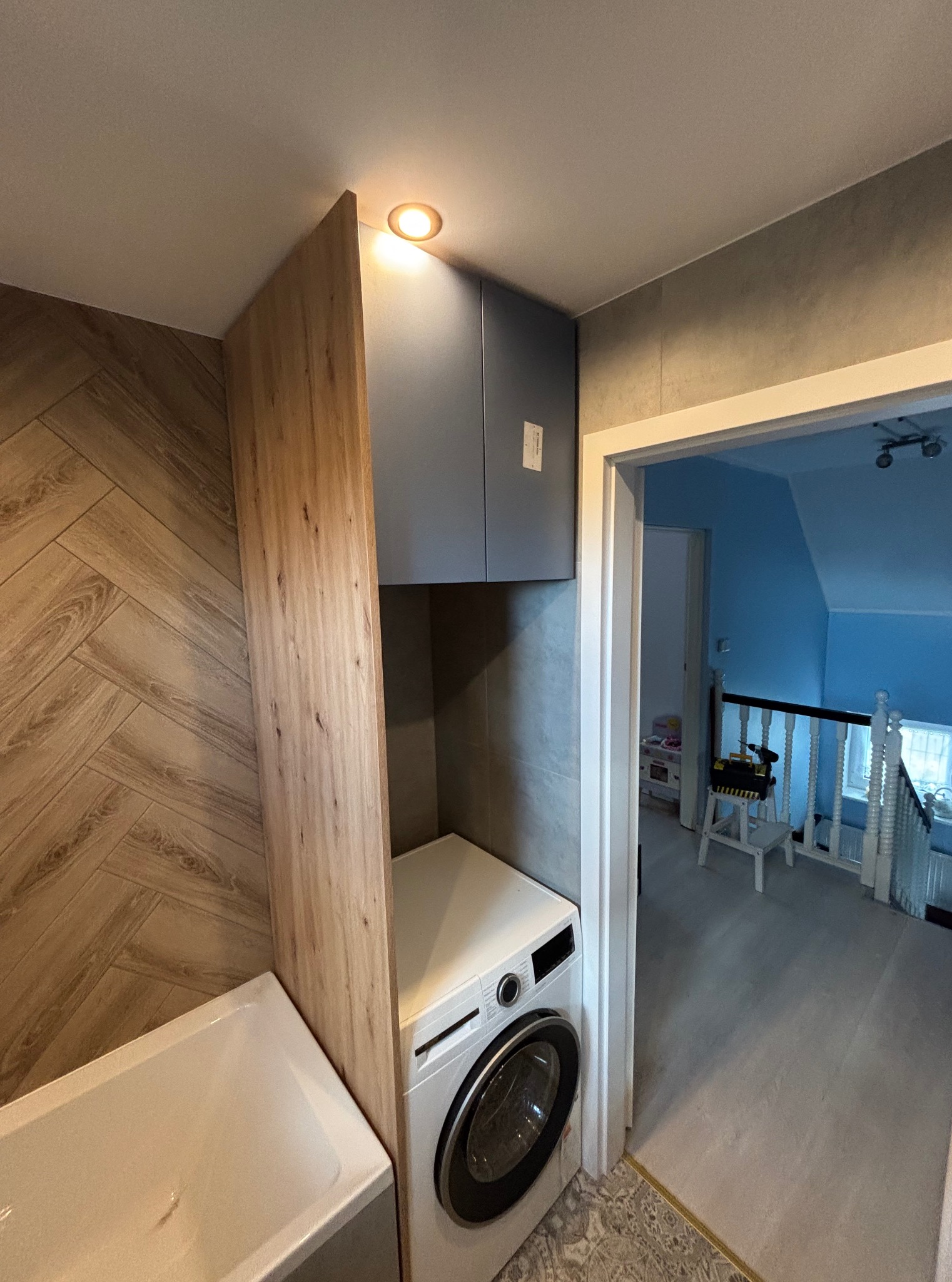 Łazienka z zabudową meblową: szafka nad pralką, drewniana obudowa wanny, płytki imitujące drewno na ścianie. Widok na korytarz z zabawkami w tle.