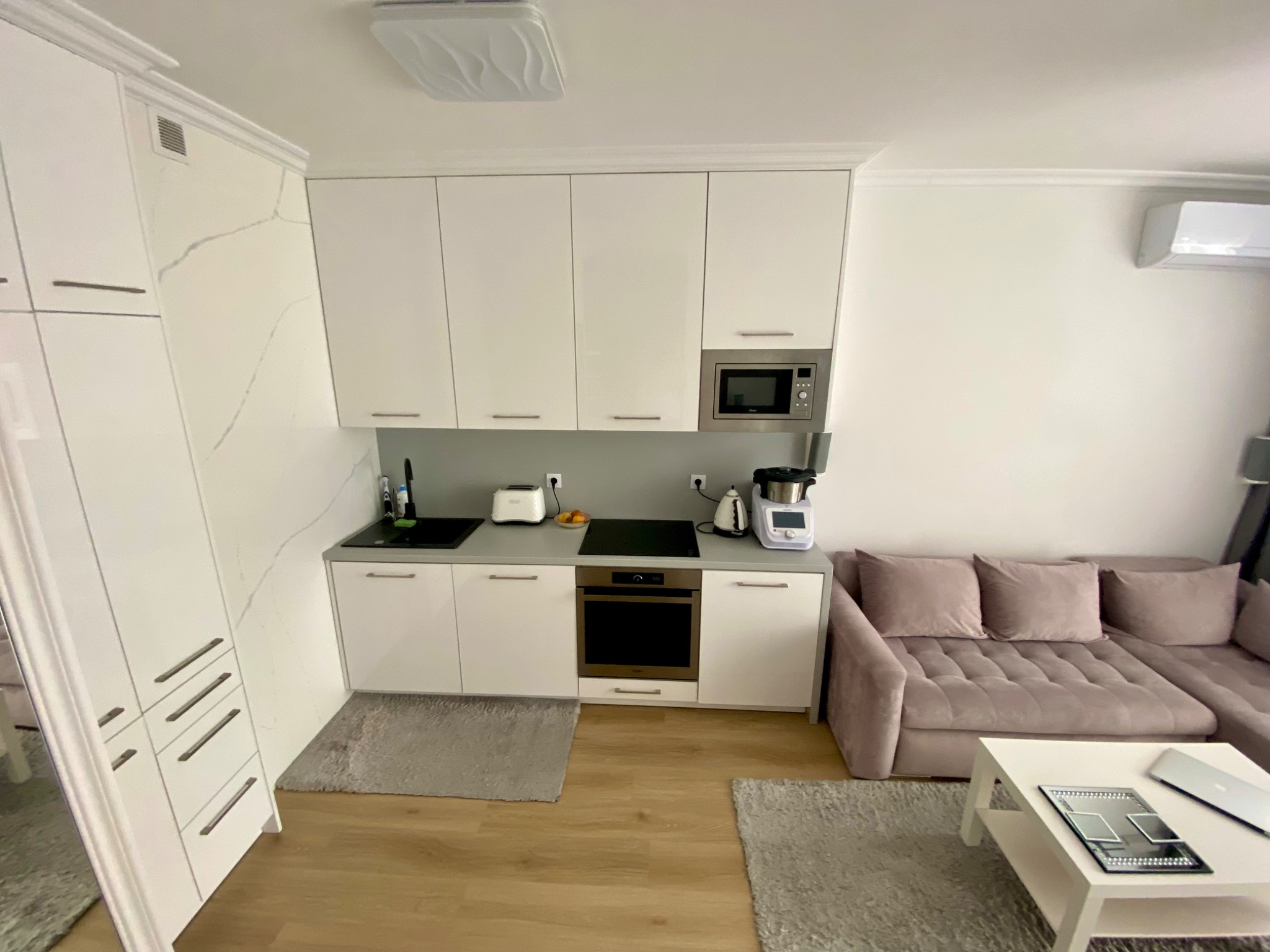 Biała zabudowa kuchenna z szafkami wiszącymi i sprzętem AGD, widok z góry na salon z sofą w odcieniach różu i stolikiem kawowym. Nowoczesne wykończenie.