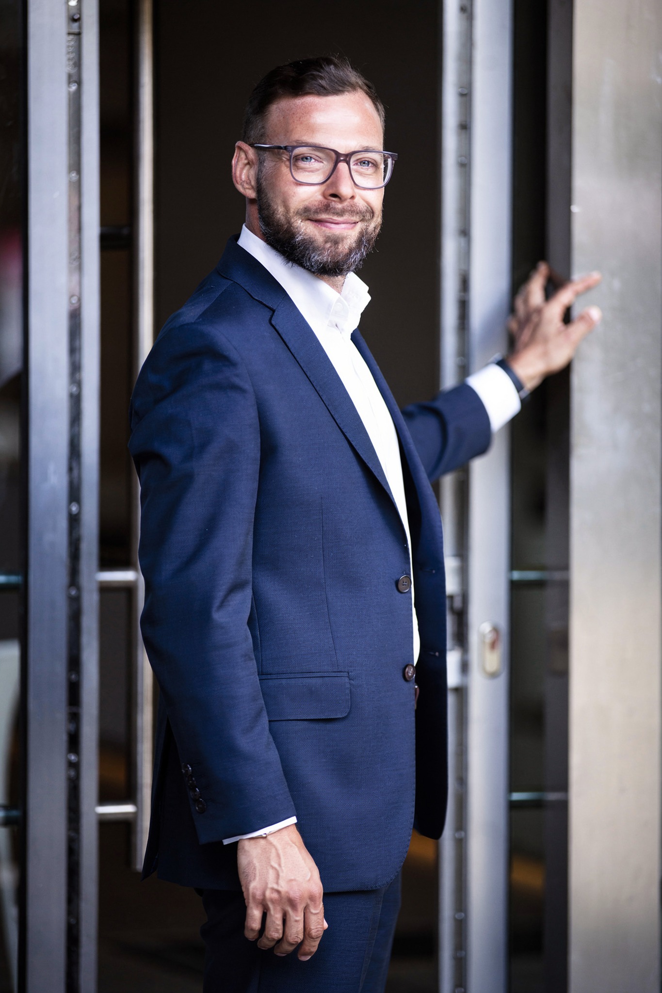 Uśmiechnięty mężczyzna w garniturze i okularach, stojący przy metalowych drzwiach. Profesjonalny wizerunek agenta nieruchomości, budzący zaufanie.