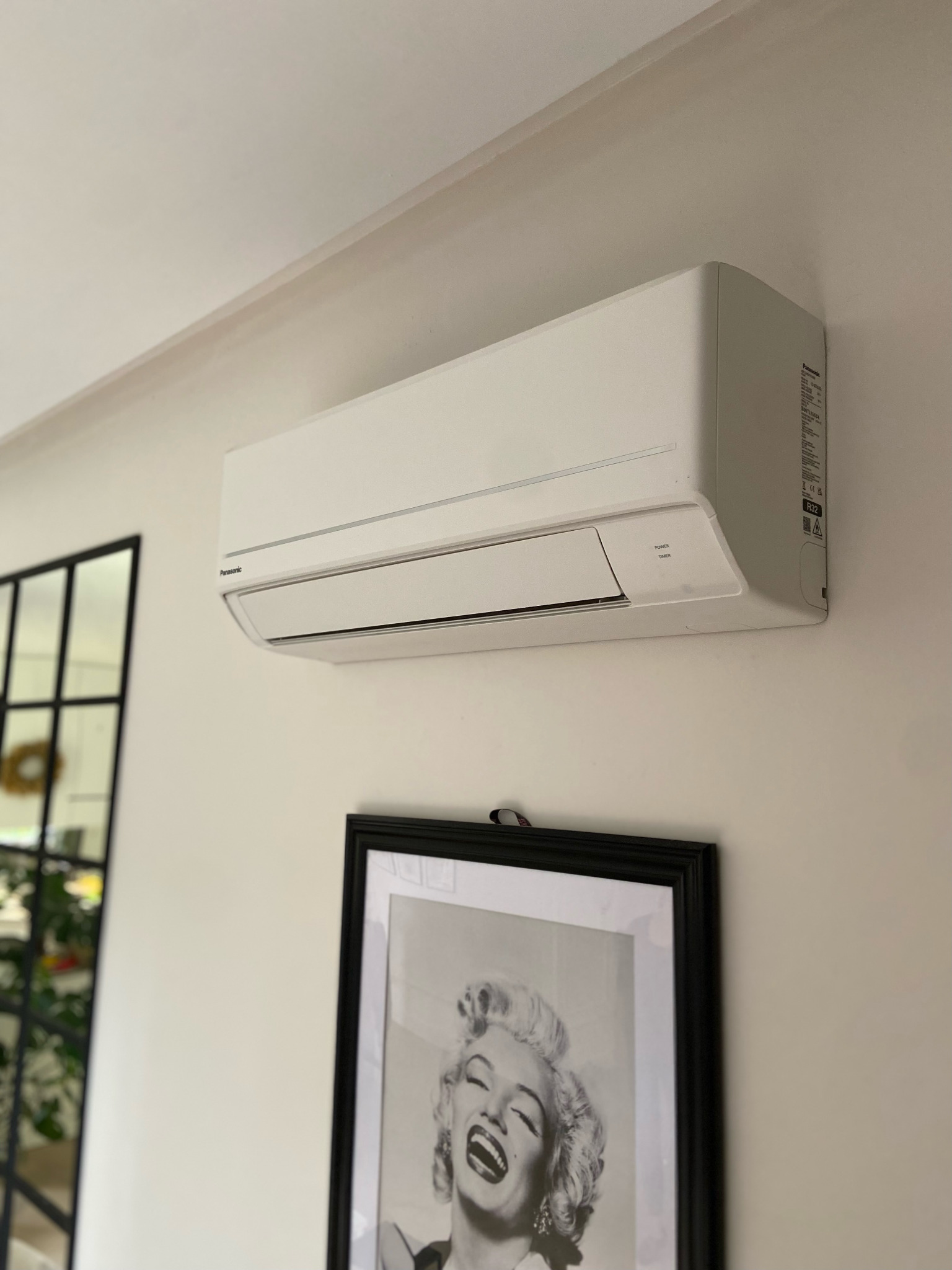 Biała klimatyzacja Panasonic zamontowana na ścianie w Grodzisku Mazowieckim. Pod spodem czarno-biała fotografia w ramie. Widoczne elementy dekoracyjne.