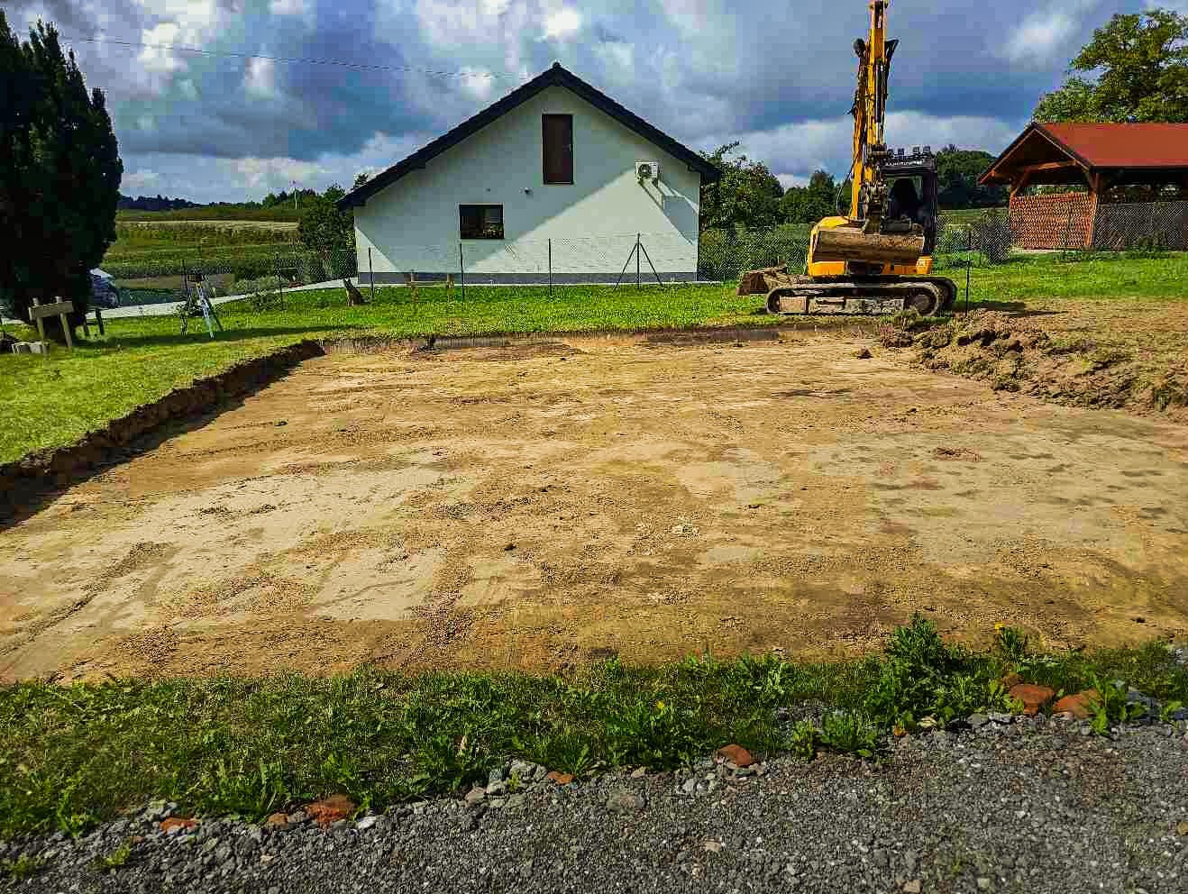 Przygotowany teren pod budowę domu, z wykopem pod fundamenty i żółtą koparką na działce. Widoczny dom w tle i fragment ogrodu.