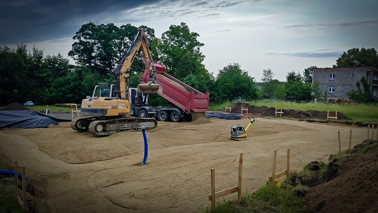 Żółta koparka Liebherr 313 wysypuje piasek z czerwonej wywrotki na przygotowany teren pod budowę, obok zagęszczarka. Widoczne ogrodzenie i budynek w tle.