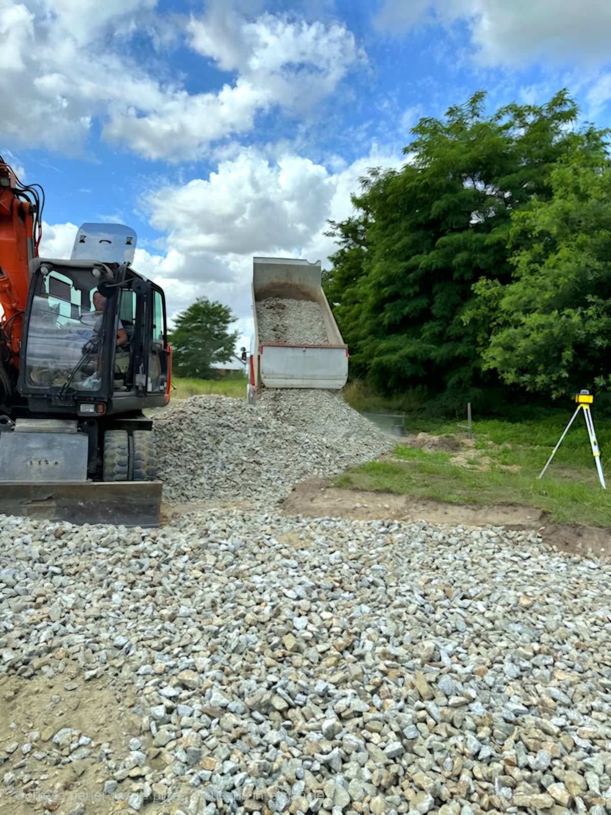 Pomarańczowa koparka i wywrotka wysypująca żwir na teren. W tle zielone drzewa i błękitne niebo z chmurami. Widoczny niwelator laserowy na statywie.