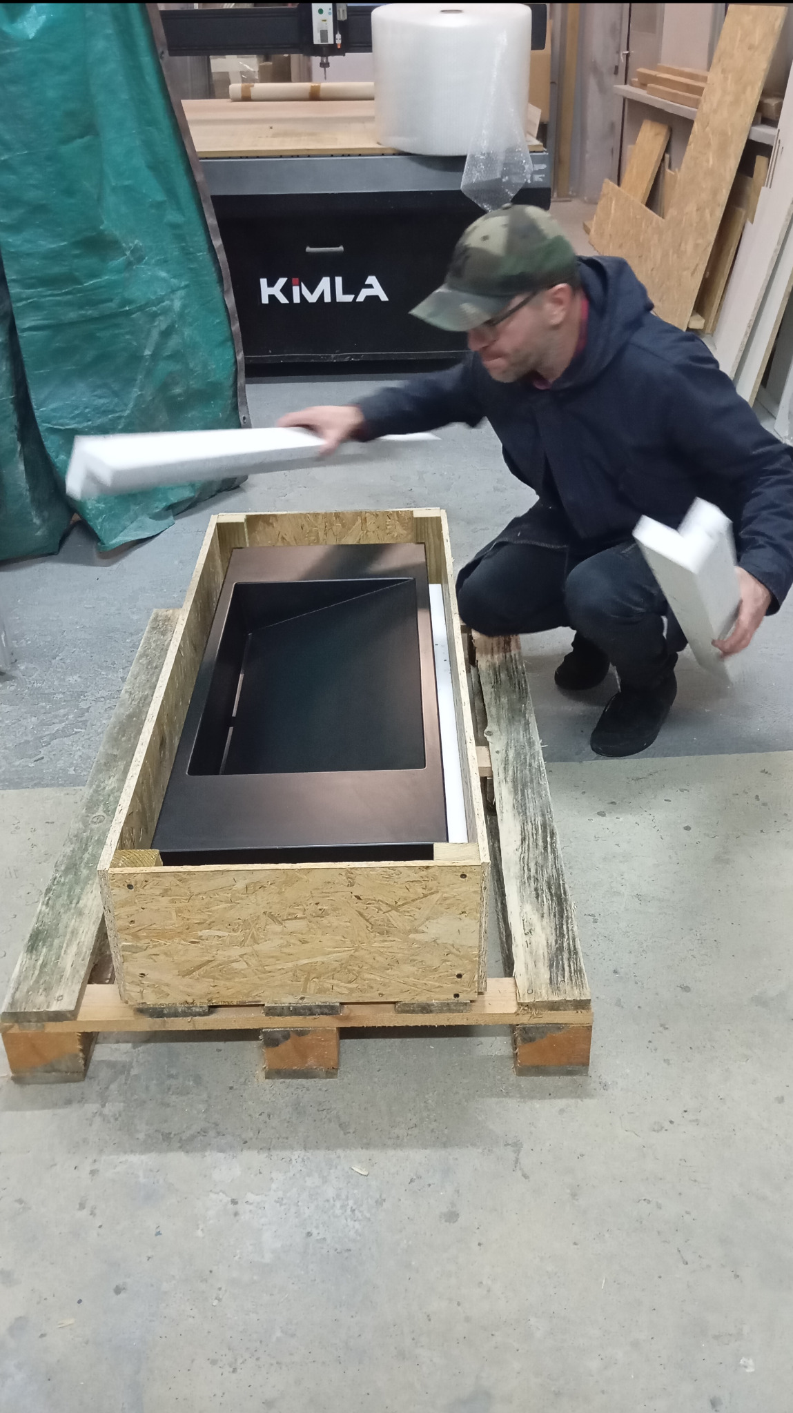 Czarny prefabrykat w drewnianej skrzyni, zabezpieczany styropianem przez mężczyznę w czapce. W tle maszyna KIMLA i materiały budowlane.