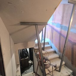 Dud-Bak - Montaż stelażu pod płytę G-K na schodach. Widoczne profile, przewody i narzędzia. Etap prac wykończeniowych wewnątrz budynku. Ujęcie z dołu schodów.