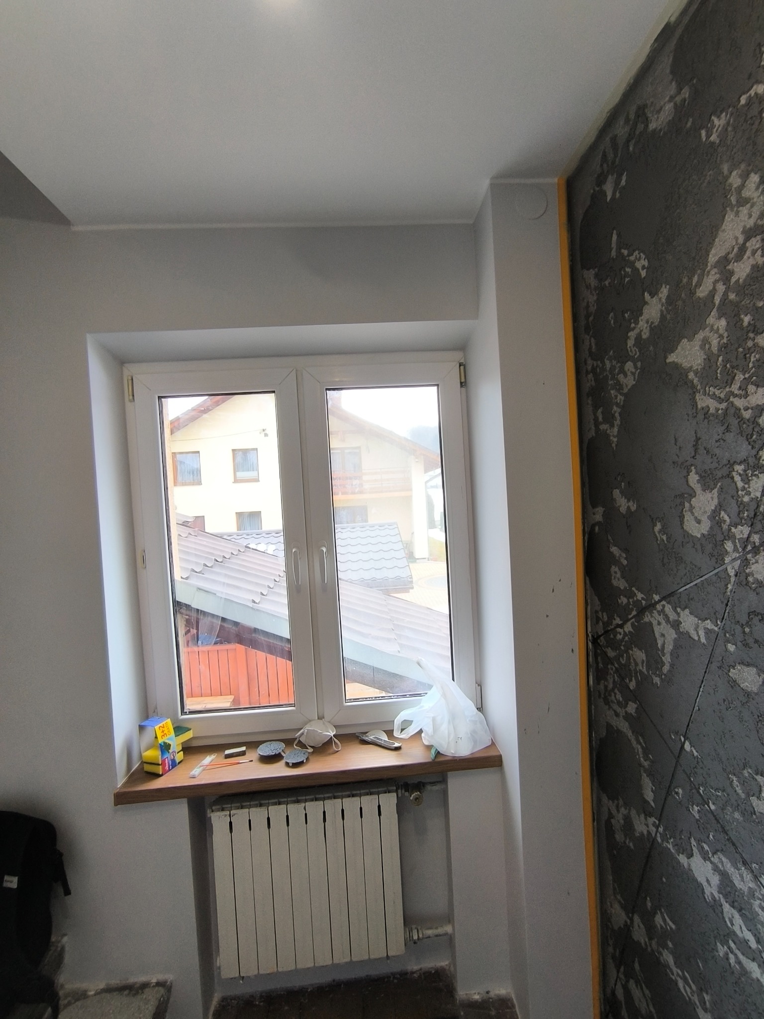 Wykończone wnętrze: okno PCV z parapetem, grzejnik, ściana z dekoracyjną strukturą. Widoczne narzędzia malarskie i akcesoria na parapecie. Szare ściany.