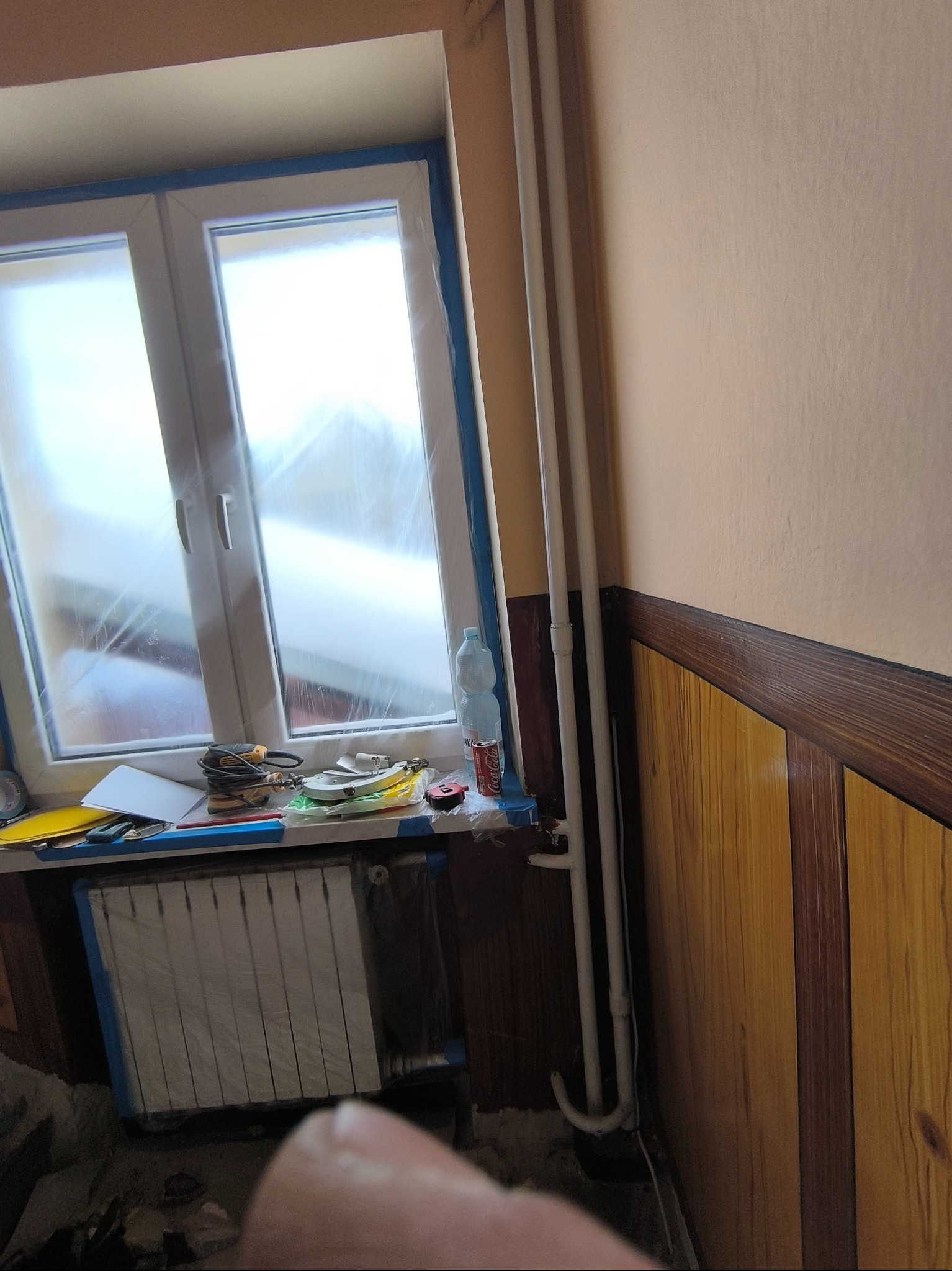 Pokój w trakcie prac malarskich: okno i grzejnik zabezpieczone folią, narzędzia na parapecie, ściana w kolorze brzoskwini, drewniana boazeria. Przygotowanie do malowania.