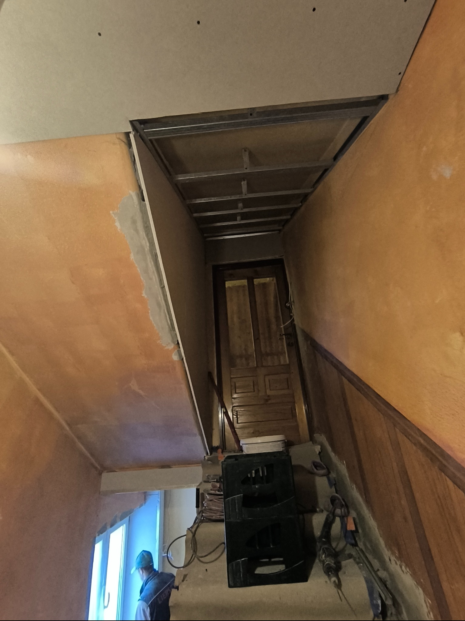 Sufit z płyt karton-gips, widoczne profile metalowe i częściowo wyszpachlowana ściana w ciepłych odcieniach. W tle drzwi i okno z pracownikiem w niebieskiej czapce.