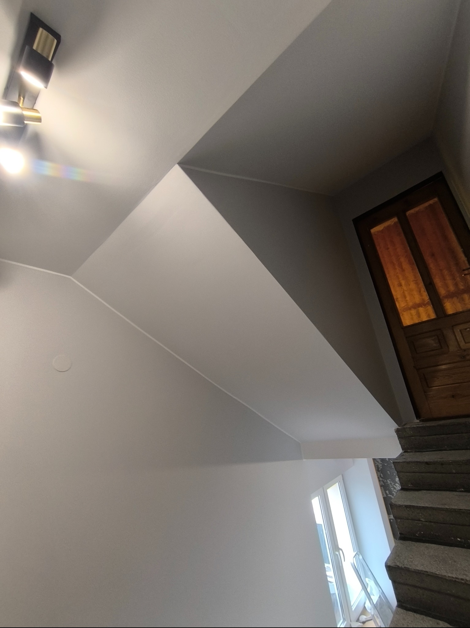 Świeżo pomalowana klatka schodowa w szarościach, z widocznymi schodami, oknem i drewnianymi drzwiami. Nowoczesne oświetlenie sufitowe.