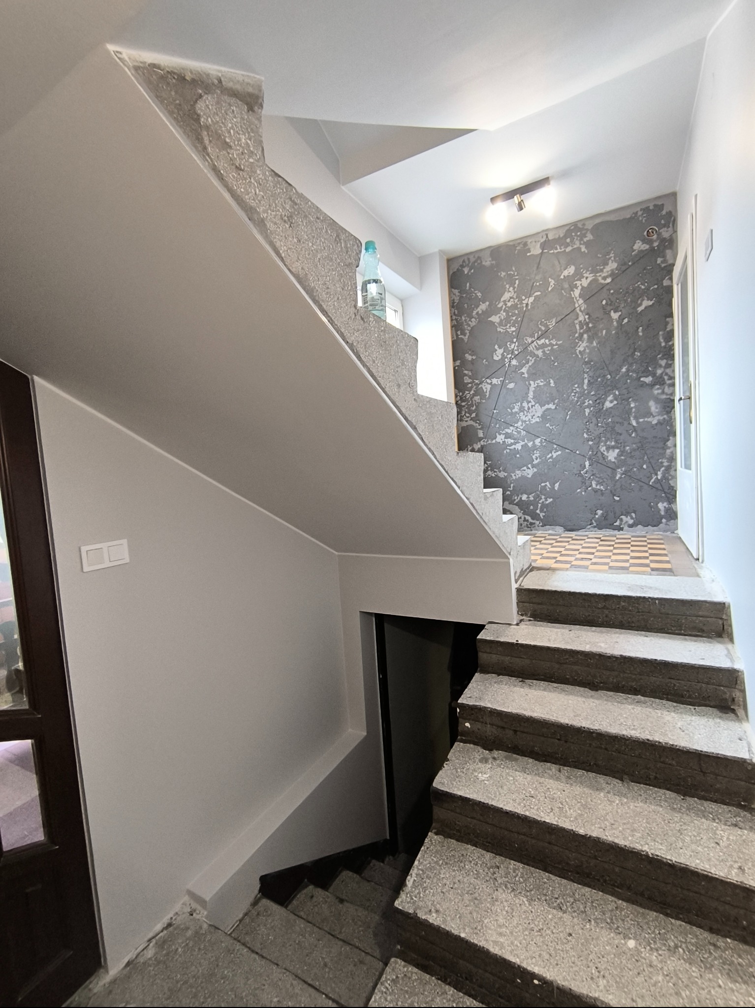 Wykończone schody z szarą, dekoracyjną ścianą i białymi ścianami. Widoczna butelka wody na schodach. Minimalistyczny design.