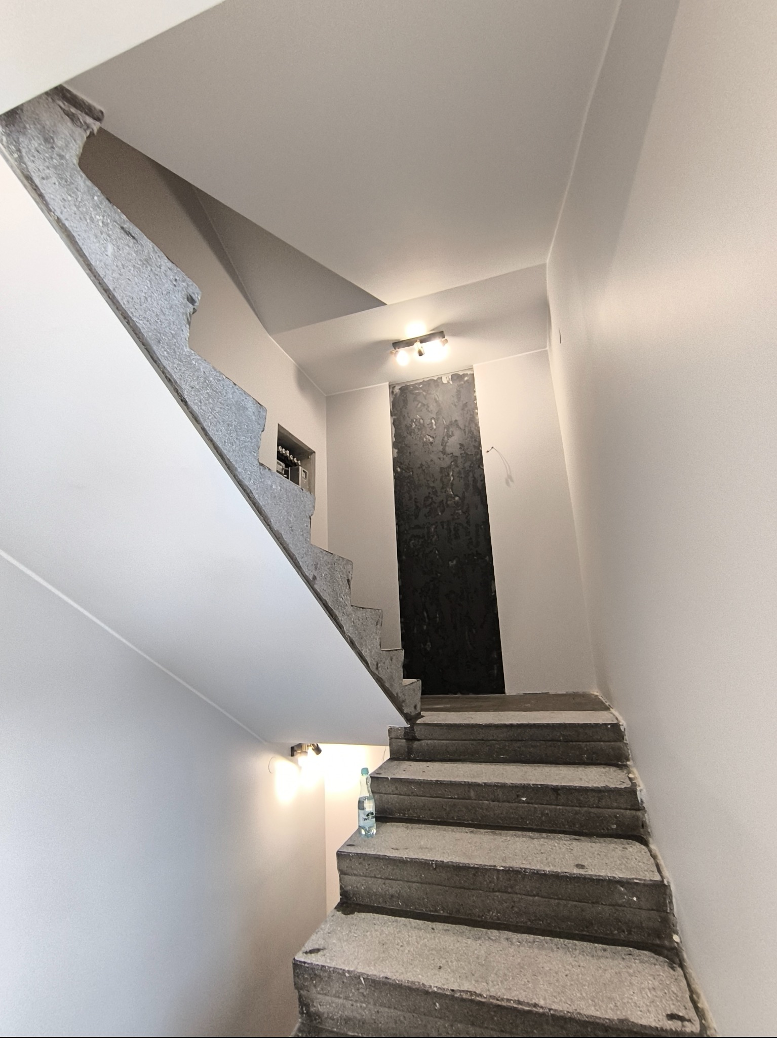 Klatka schodowa w trakcie remontu: surowe betonowe schody, świeżo pomalowane ściany na biało, widoczne niedoskonałości i przygotowania do dalszych prac wykończeniowych.