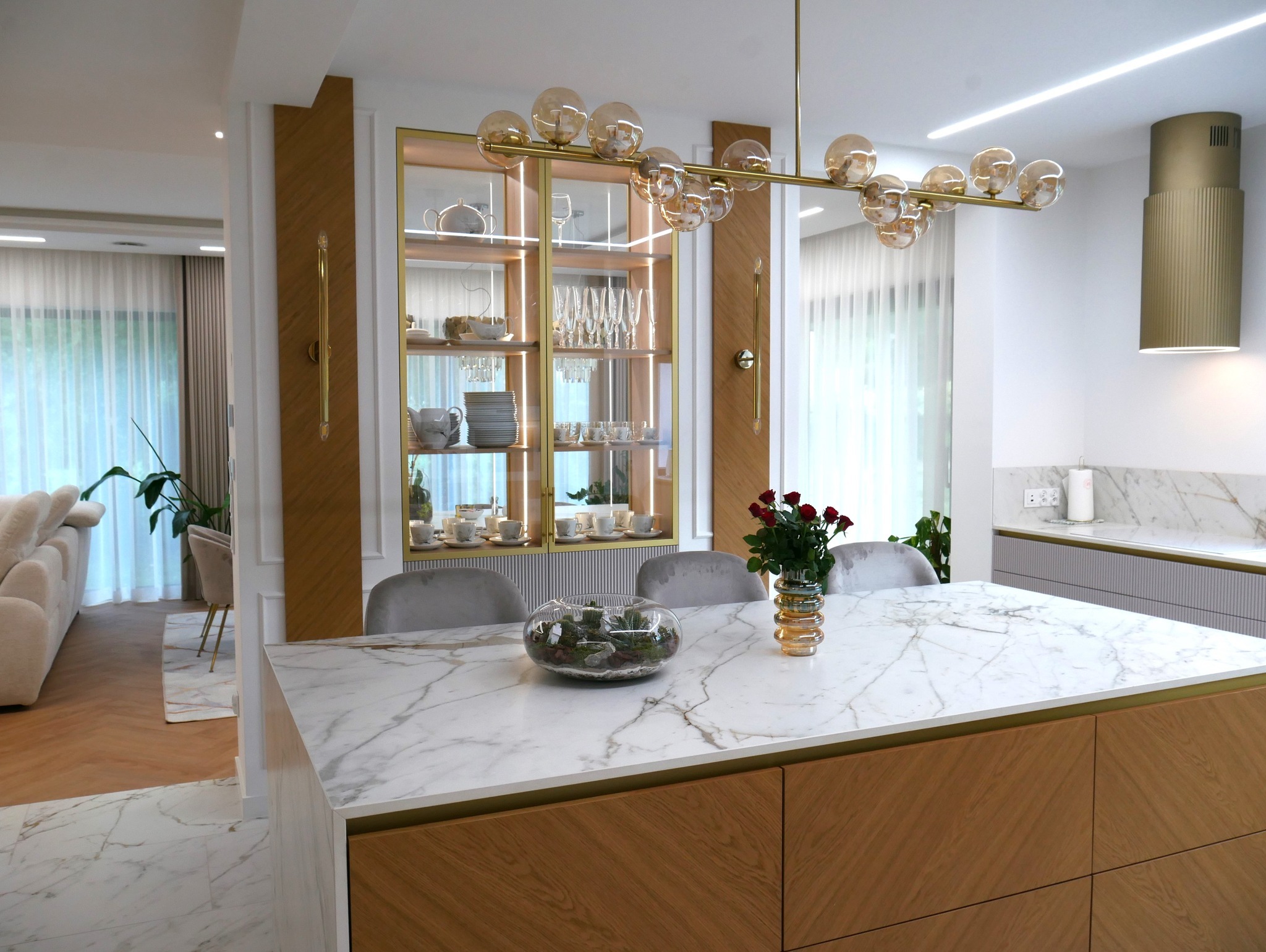 Elegancka kuchnia z marmurowym blatem wyspy, złotymi akcentami i przeszkloną witryną. Wazon z różami i szklana kula z roślinami dodają wnętrzu przytulności.