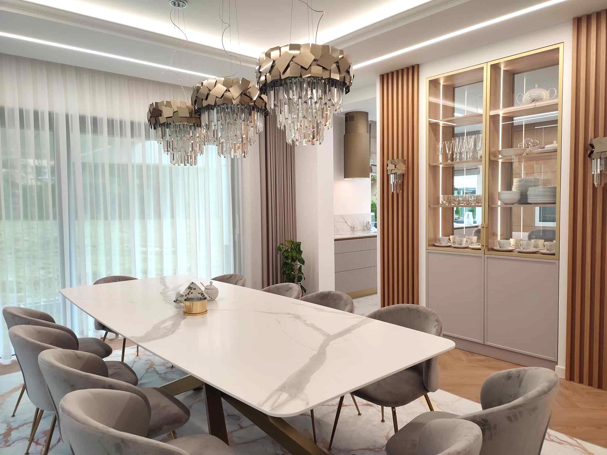 Elegancka jadalnia z marmurowym stołem, szarymi krzesłami i kryształowymi żyrandolami. W tle przeszklona witryna z zastawą i widok na kuchnię.