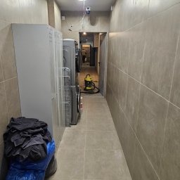 Michał partyga - Wykafelkowane, wąskie pomieszczenie z pralkami i workami na podłodze. W tle widoczny odkurzacz i osoba w przejściu. Stonowana kolorystyka płytek.