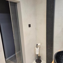 Michał partyga - Wykończona łazienka z jasnymi płytkami, czarnym panelem dekoracyjnym i elementami wyposażenia w ciemnych kolorach. Widoczny fragment kabiny prysznicowej i umywalki.