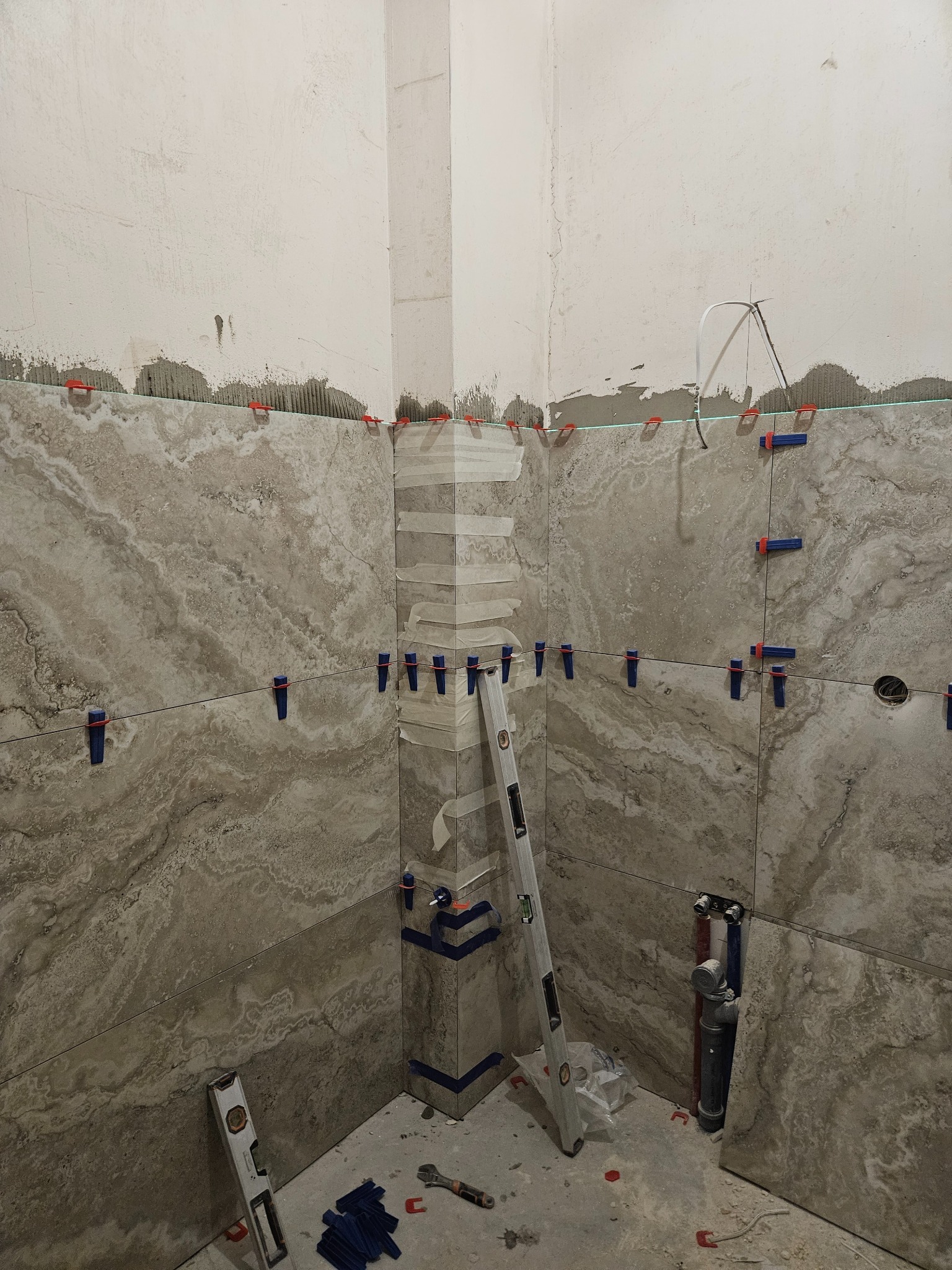 Układanie płytek imitujących marmur w rogu łazienki, widoczne poziomice, krzyżyki dystansowe i fragment instalacji wodnej. Prace w toku, surowy stan ścian.
