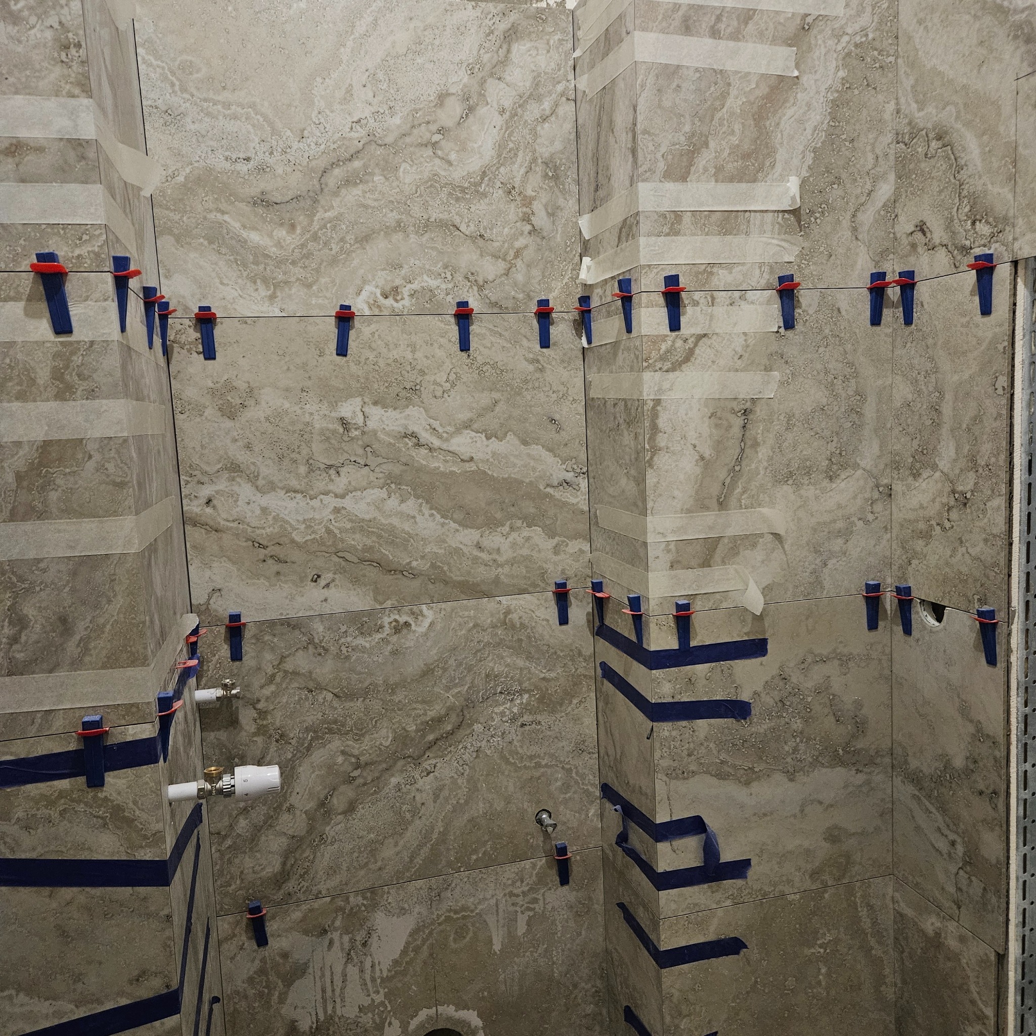 Ściana w trakcie układania płytek imitujących marmur, z użyciem poziomowania klinami i taśmy. Widoczne fragmenty instalacji wodnej i metalowy profil narożny.