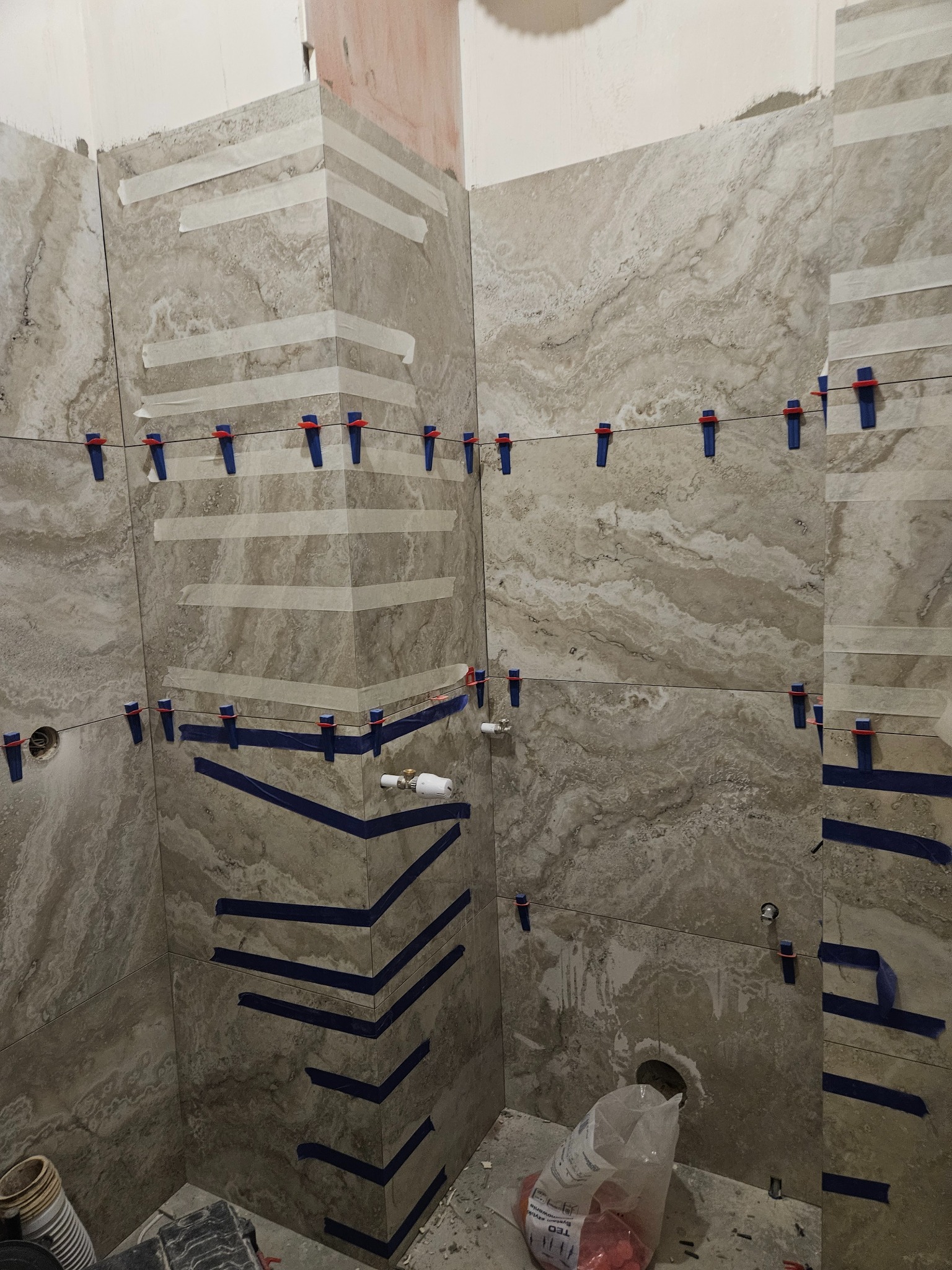 Układanie płytek imitujących marmur w łazience, z użyciem poziomowania krzyżykami i taśmy maskującej. Widoczne otwory na instalacje i akcesoria łazienkowe.