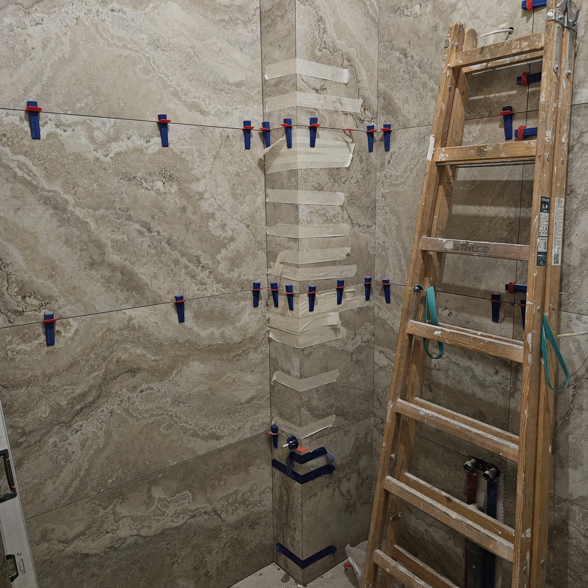 Układanie płytek w łazience z użyciem poziomowania, widoczna drabina i fragment instalacji. Płytki w odcieniach beżu z wyraźnym wzorem.