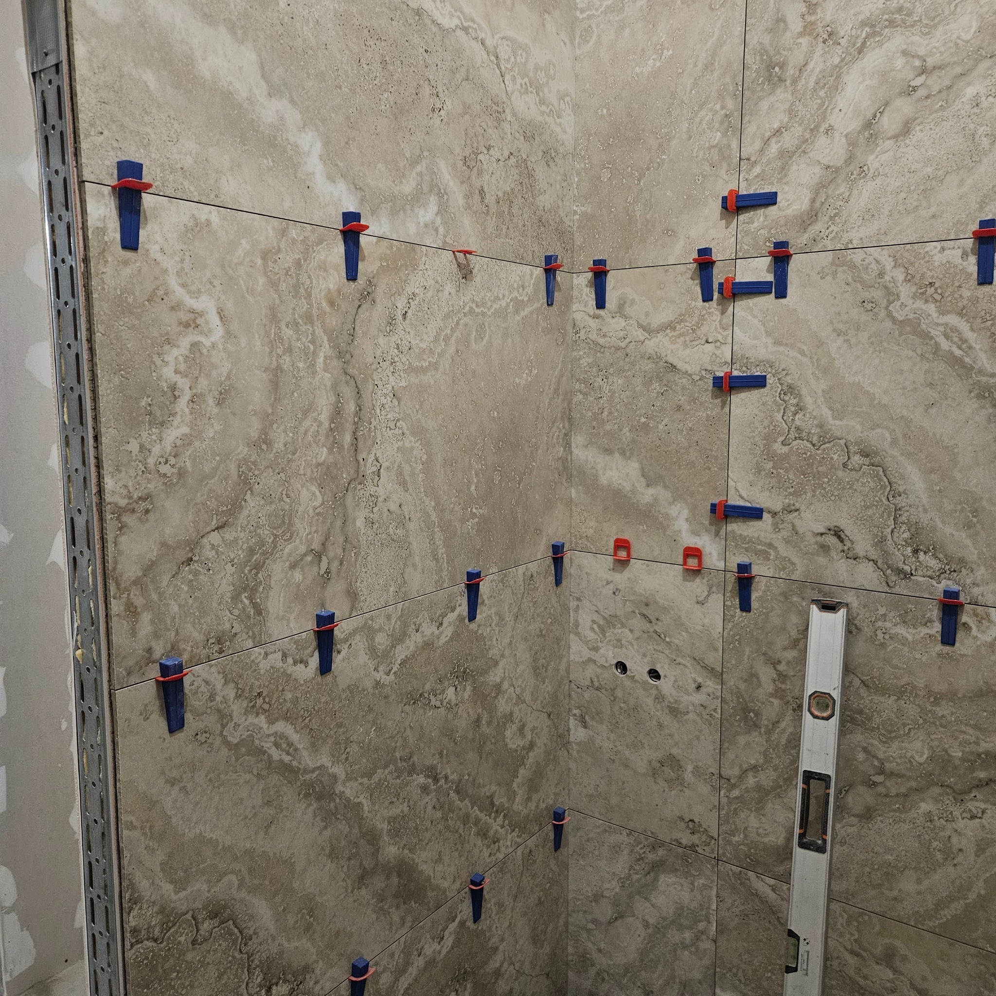 Układanie beżowych płytek imitujących marmur w rogu pomieszczenia, z użyciem poziomicy i systemu poziomowania płytek (klipsy i kliny). Widoczny profil narożny.