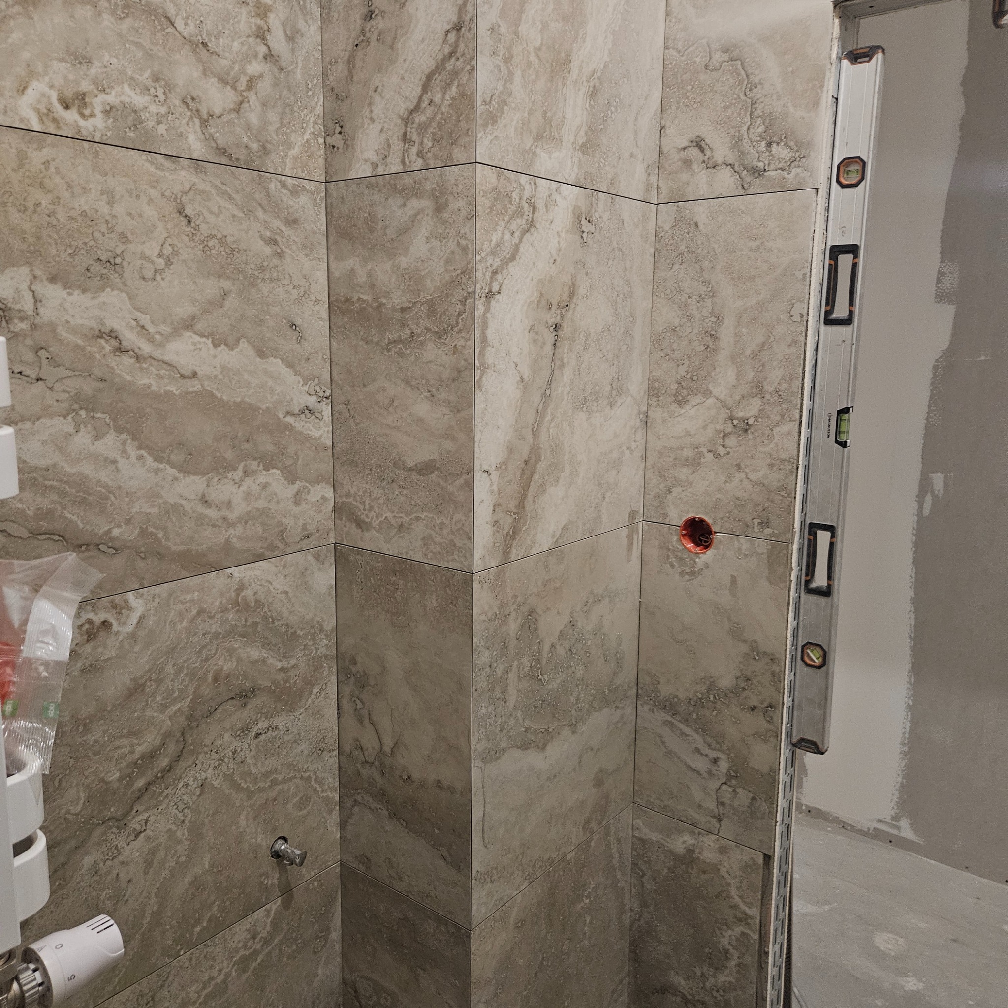 Ściana wyłożona beżowymi płytkami imitującymi marmur, z widocznym poziomowaniem przy użyciu poziomicy. W tle surowa ściana i instalacja grzewcza.