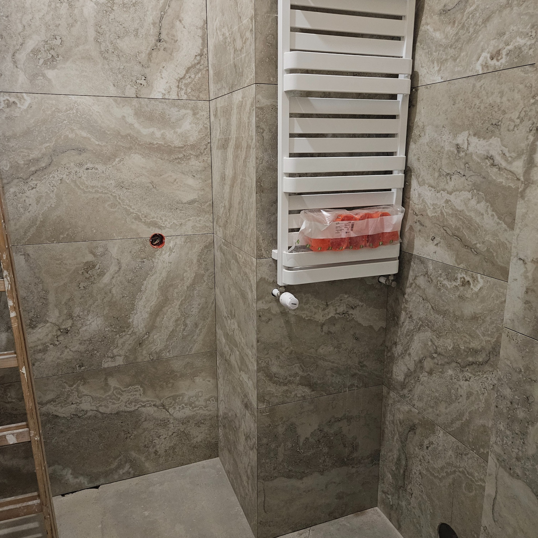 Wykończona łazienka z beżowymi płytkami imitującymi kamień, na ścianie biały grzejnik drabinkowy z termostatem. Widoczne gniazdko elektryczne i fragment drabiny.