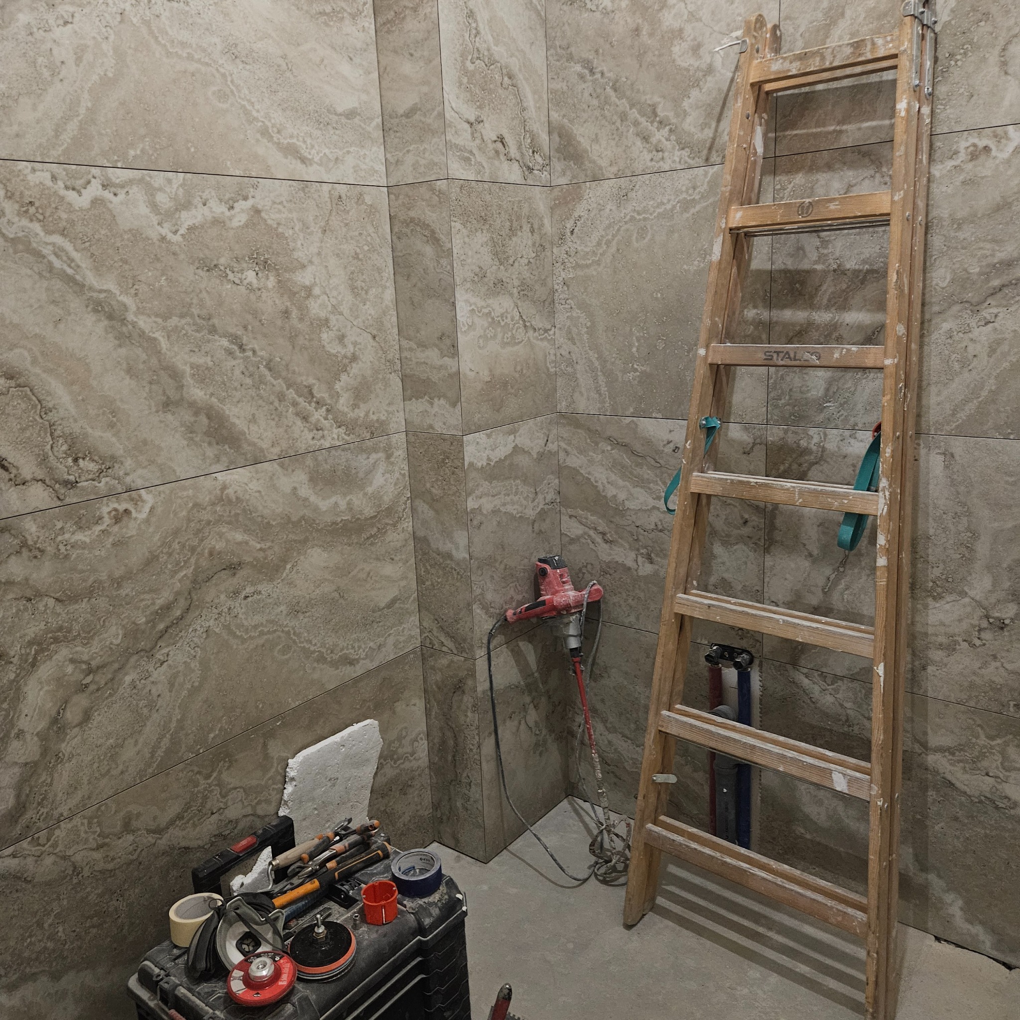 Łazienka w trakcie wykończenia: beżowe płytki imitujące marmur na ścianach, drabina, mieszadło do zaprawy i skrzynka z narzędziami glazurniczymi.