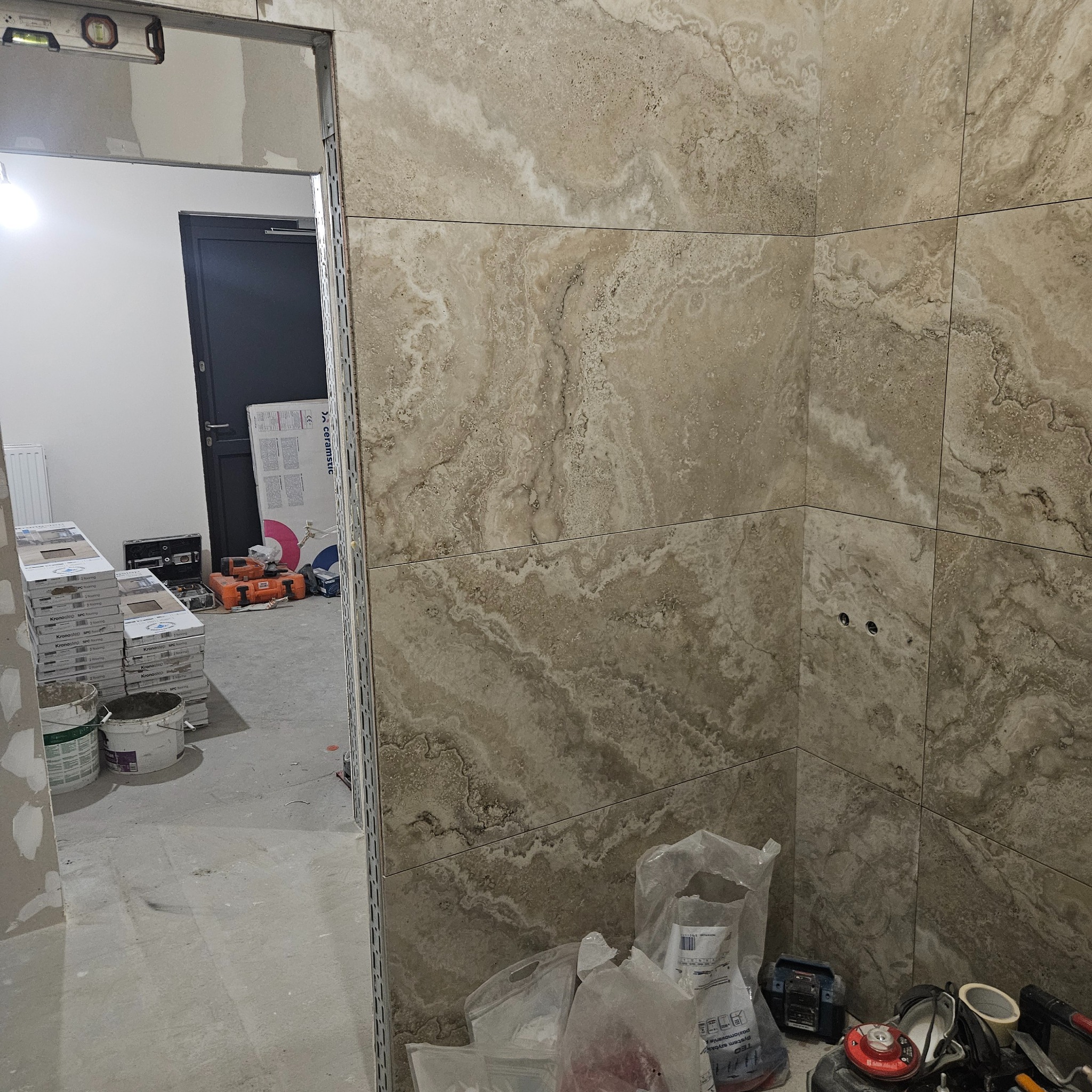 Beżowe płytki imitujące trawertyn na ścianie łazienki w trakcie wykańczania. Widoczne narzędzia i materiały budowlane. Poziomica na futrynie.