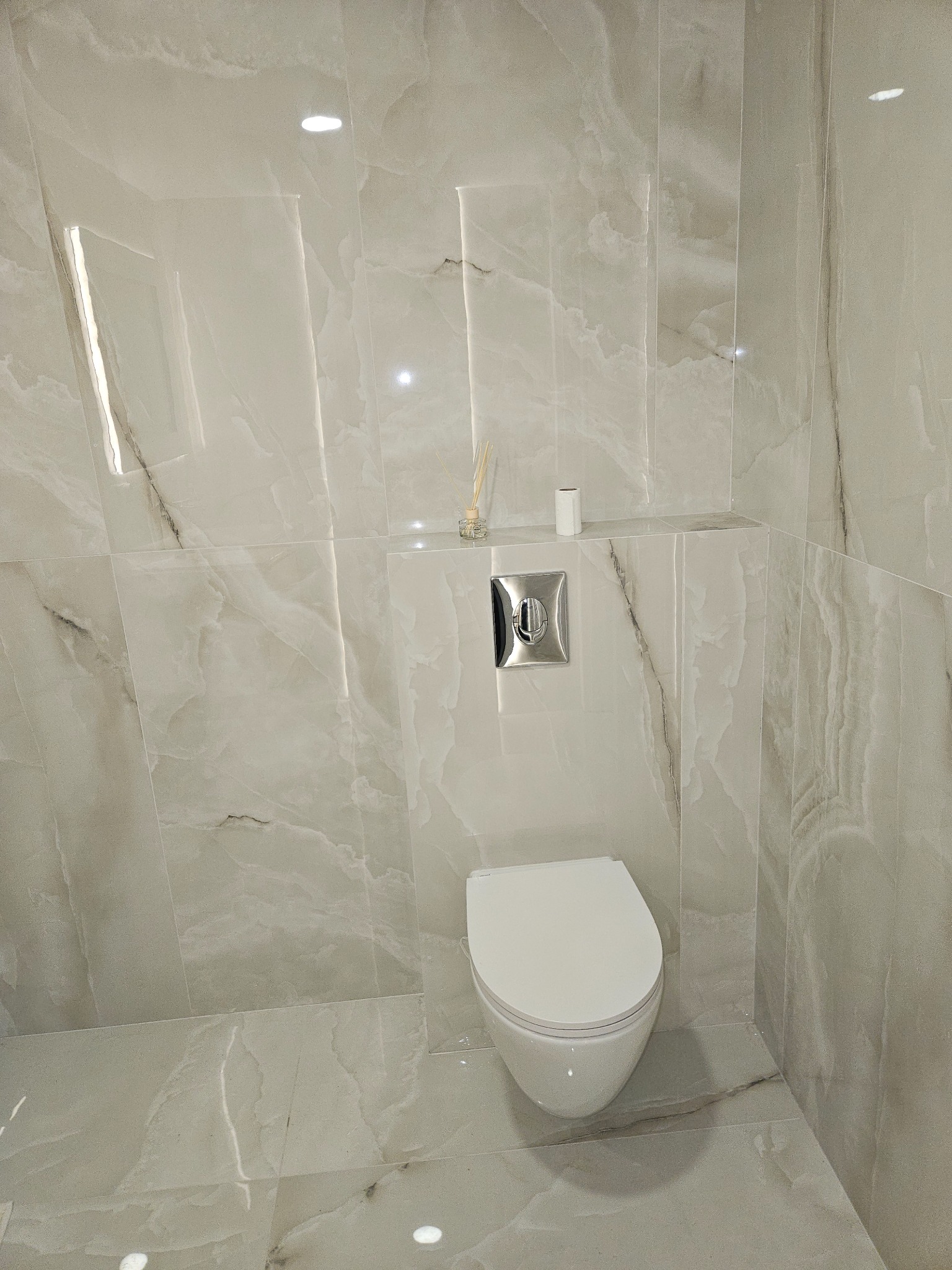 Wykończona łazienka z toaletą w odcieniach bieli i szarości. Ściany i podłoga wyłożone płytkami imitującymi marmur. Minimalistyczny design, chromowany przycisk spłuczki.