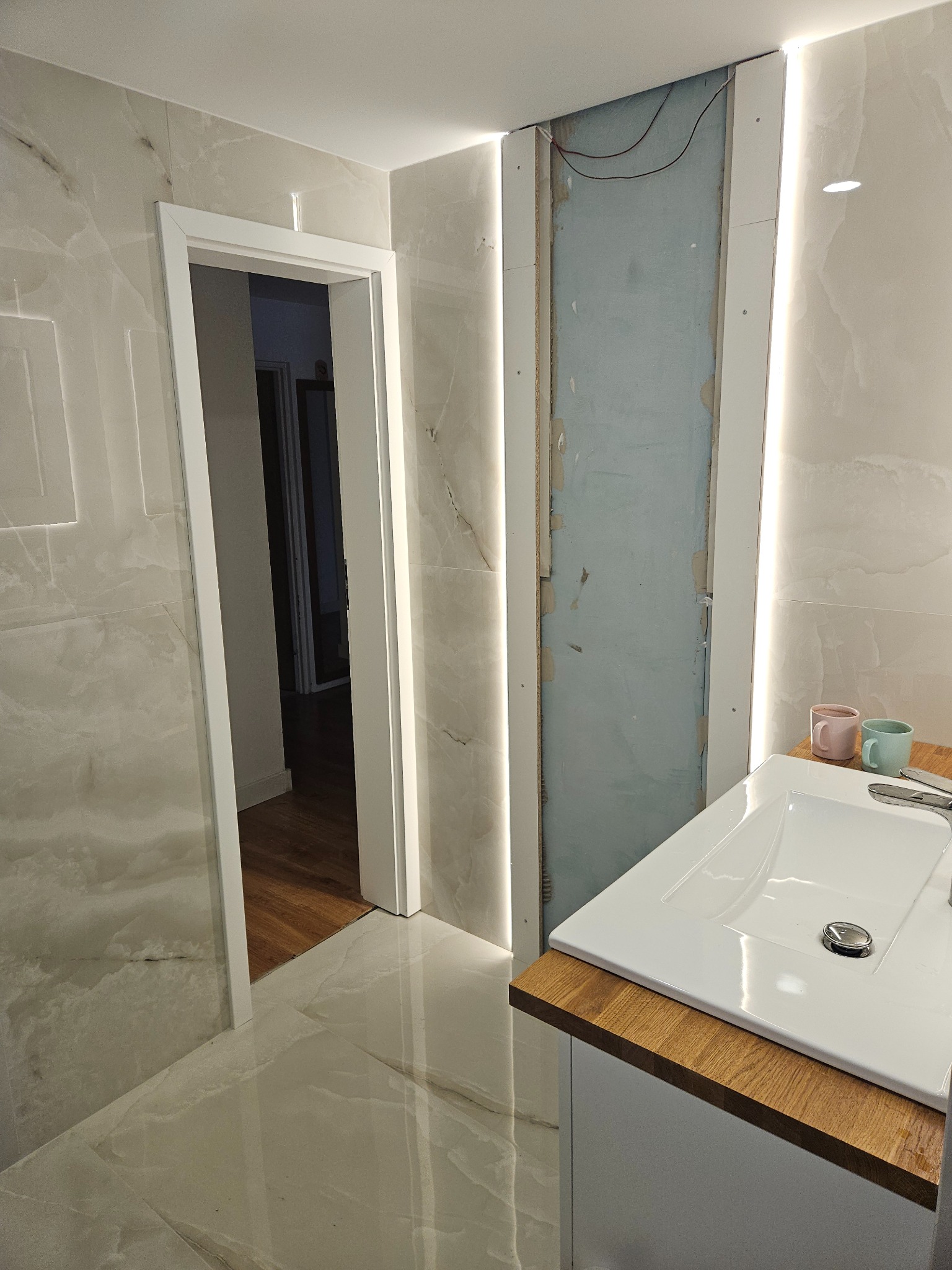 Łazienka w trakcie prac: marmurowe płytki, biała umywalka z drewnianym blatem, otwarte drzwi i fragment ściany z widoczną instalacją elektryczną. Nowoczesne oświetlenie LED.
