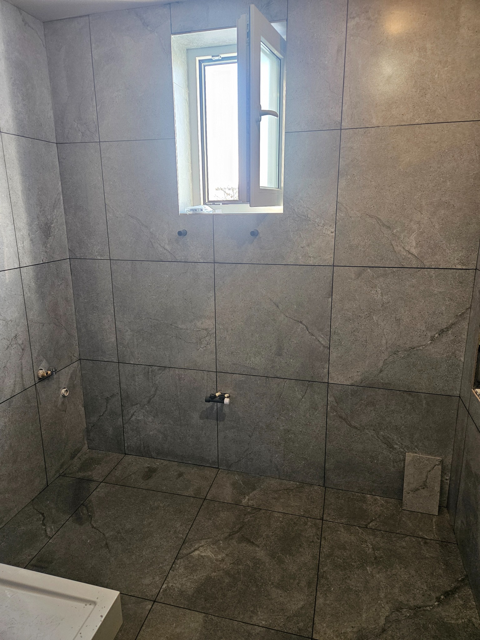 Wykończona łazienka z szarymi, kwadratowymi płytkami imitującymi kamień na ścianach i podłodze. Widoczne okno z otwartym skrzydłem i przygotowane punkty instalacji wodnej.